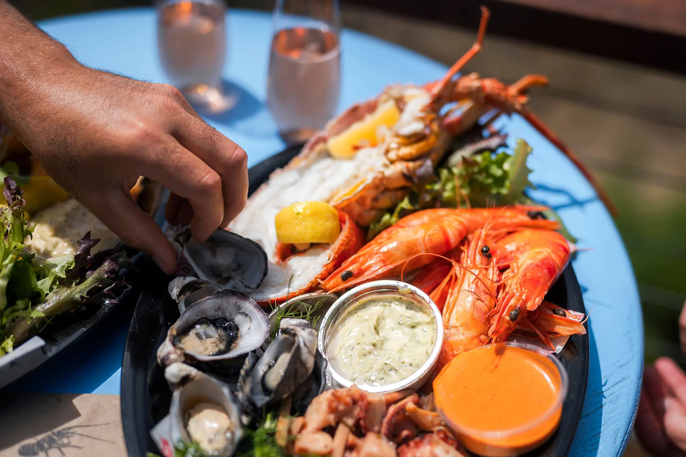A hand reaches for a seafood platter featuring lobster, oysters, lemon wedges and sauces arranged on a large platter.