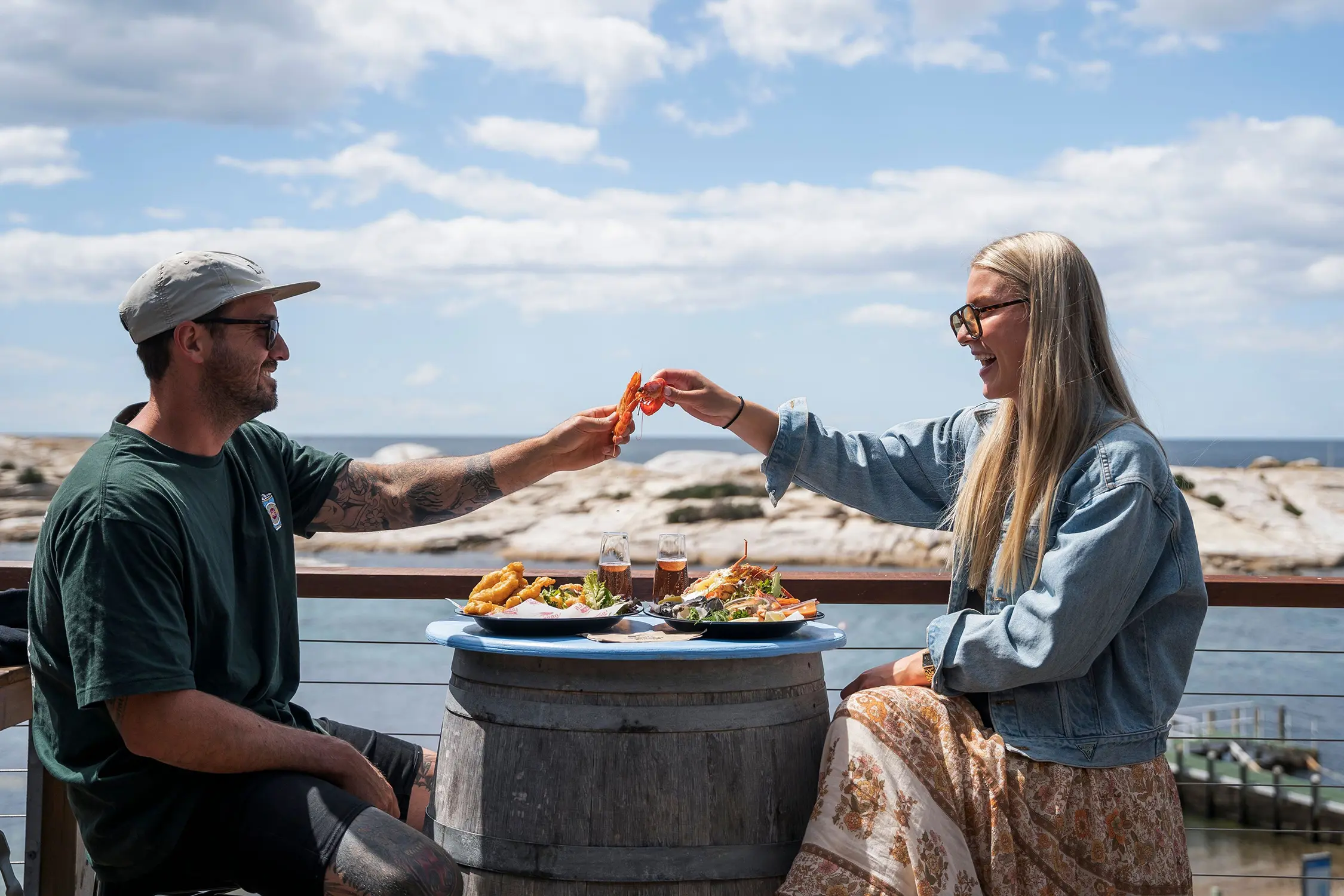 Two people sit on a seaside balcony at a repurposed wine-barrel table. The plates in front of them are loaded with fresh seafood and drinks. They share a 'toast' with a prawn each.