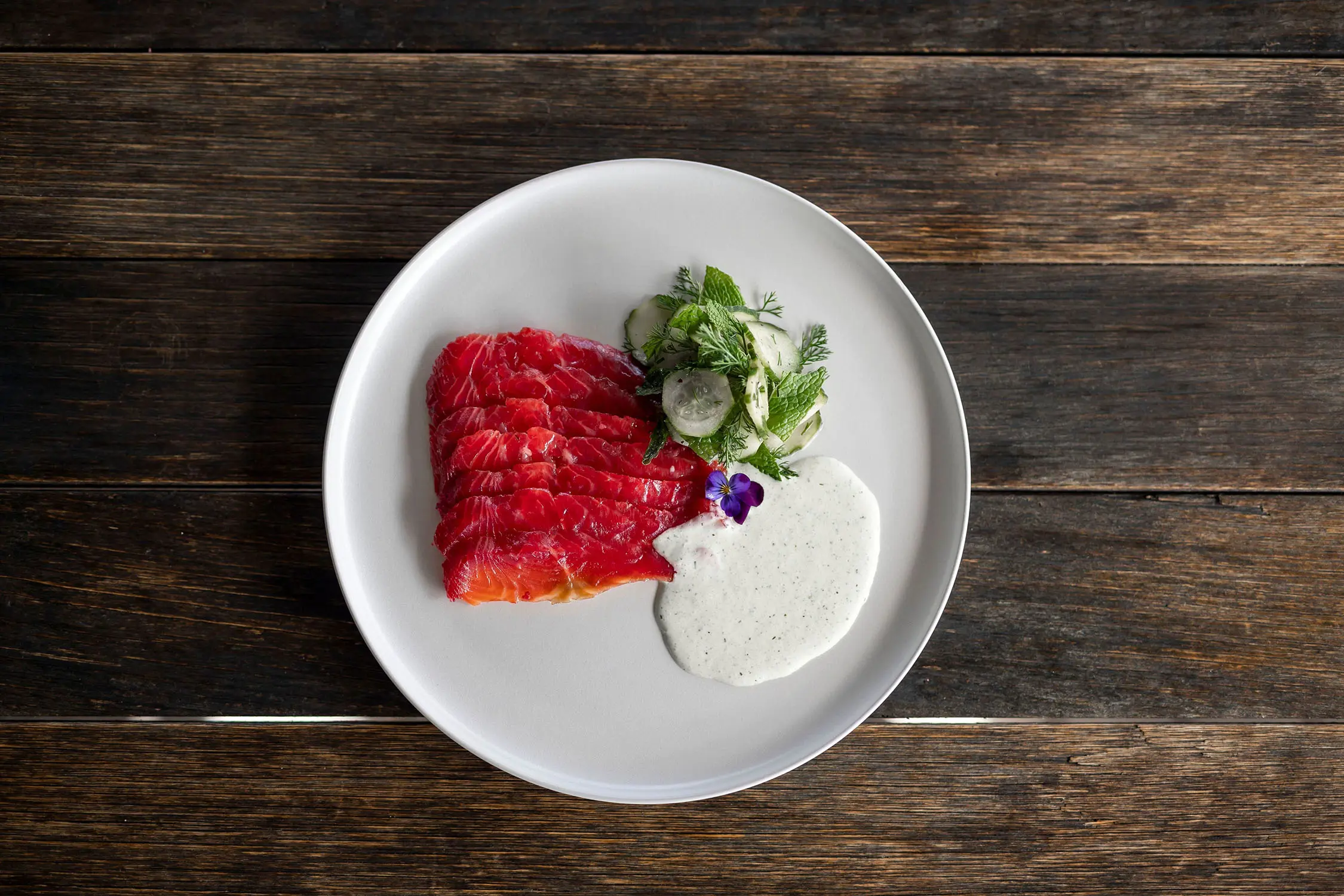 A white plate displays brightly coloured sliced salmon, with a fresh salad and creamy sauce on a rustic wooden table.