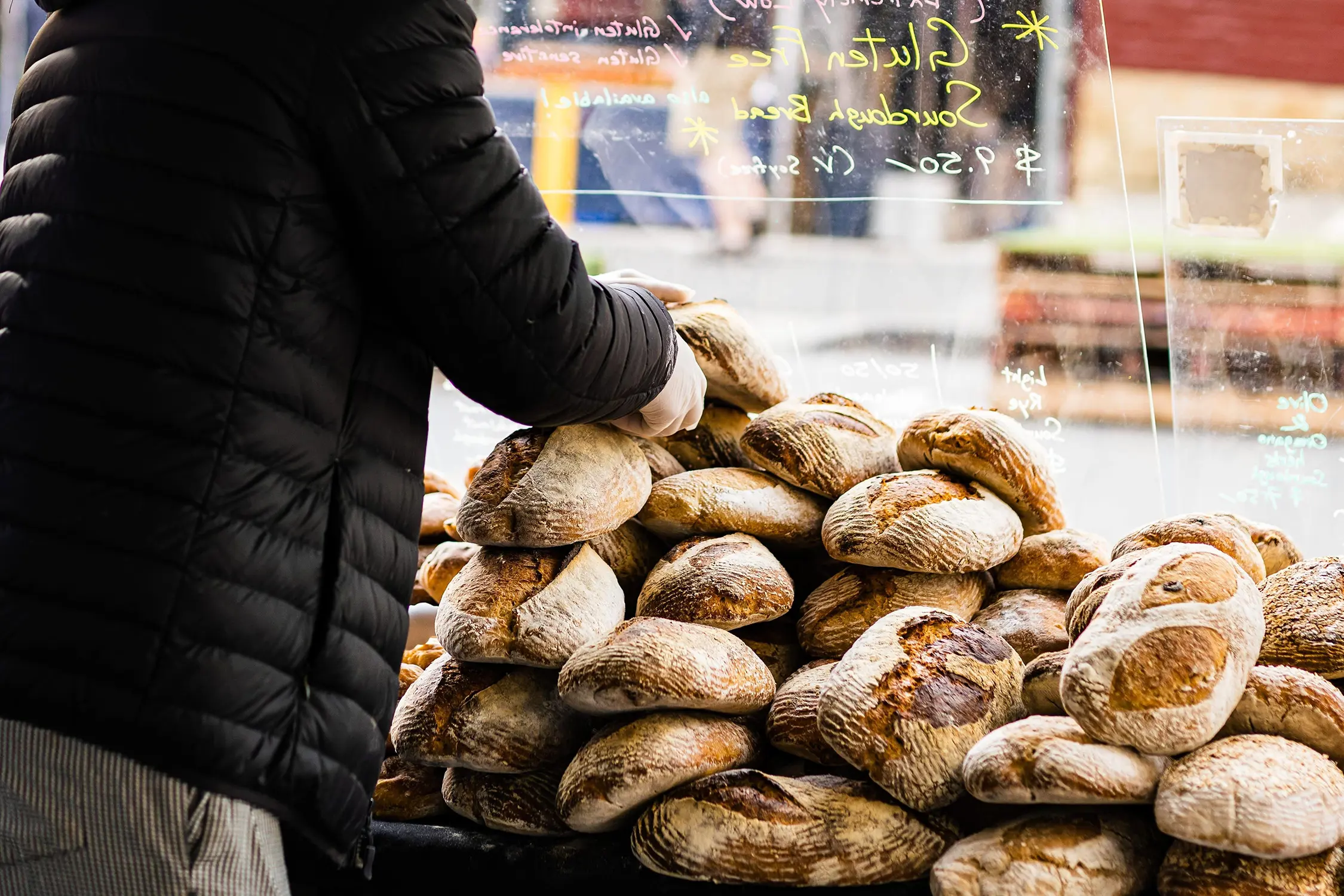 A person in a black puffer jacket stacks fresh rustic bread loaves at a market stall.