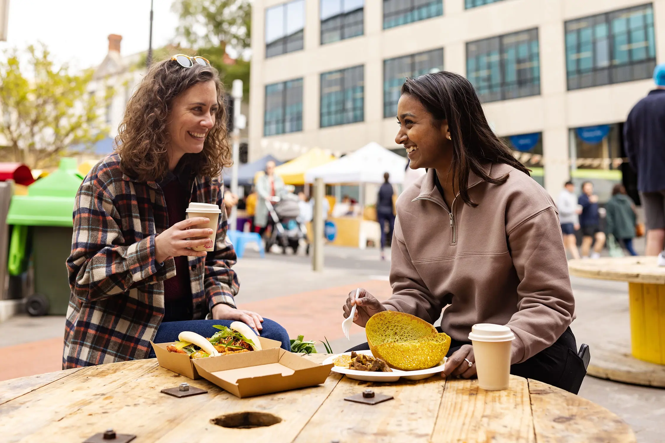 Two women sit at an outdoor table holding food and drinks, with the market crowd and buildings in background.