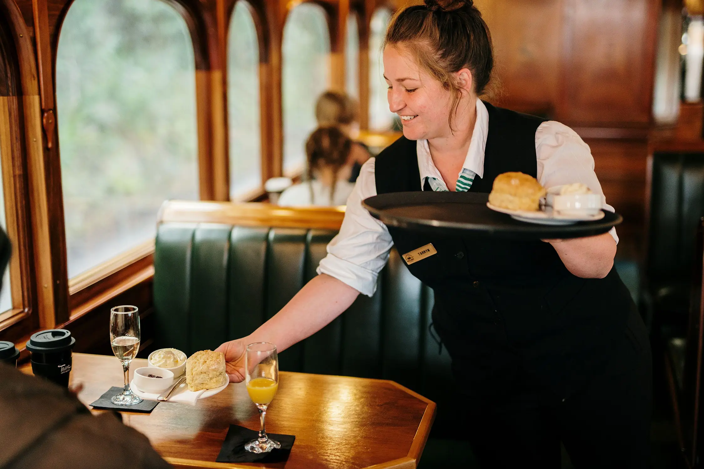 Inside a heritage train's dining car, a staff member in formal attire serves fresh scones and sparkling wine.