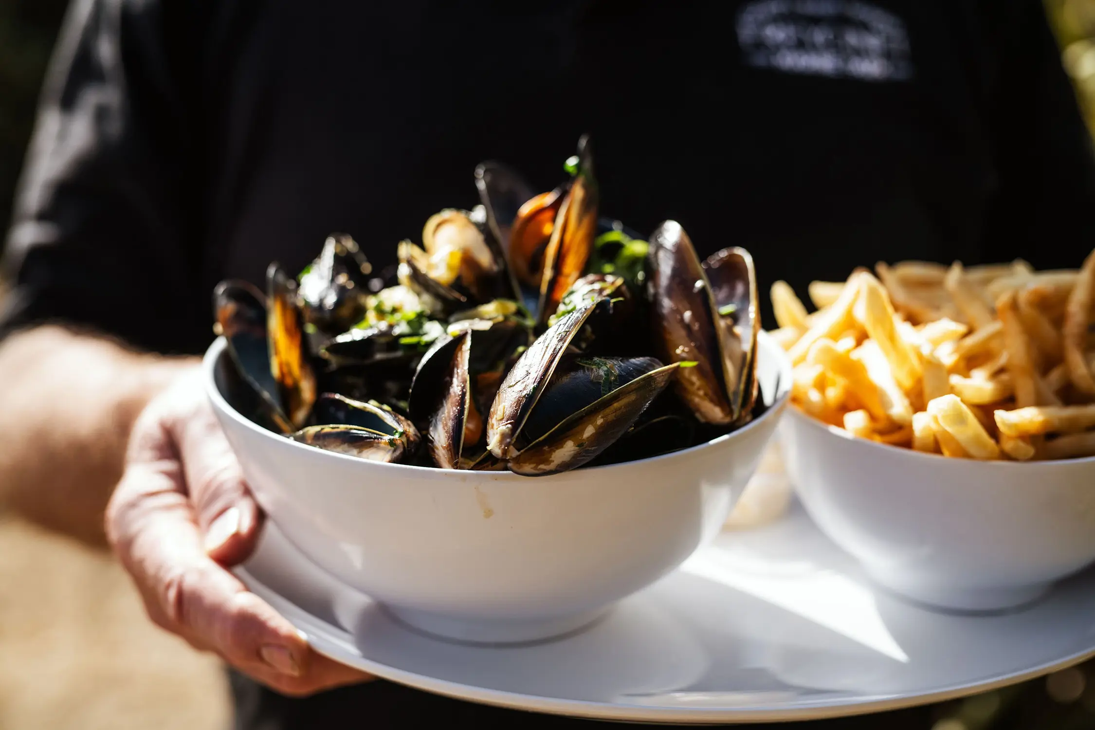 A person holds a platter with a bowl full of cooked mussels and another bowl containing crispy chips.