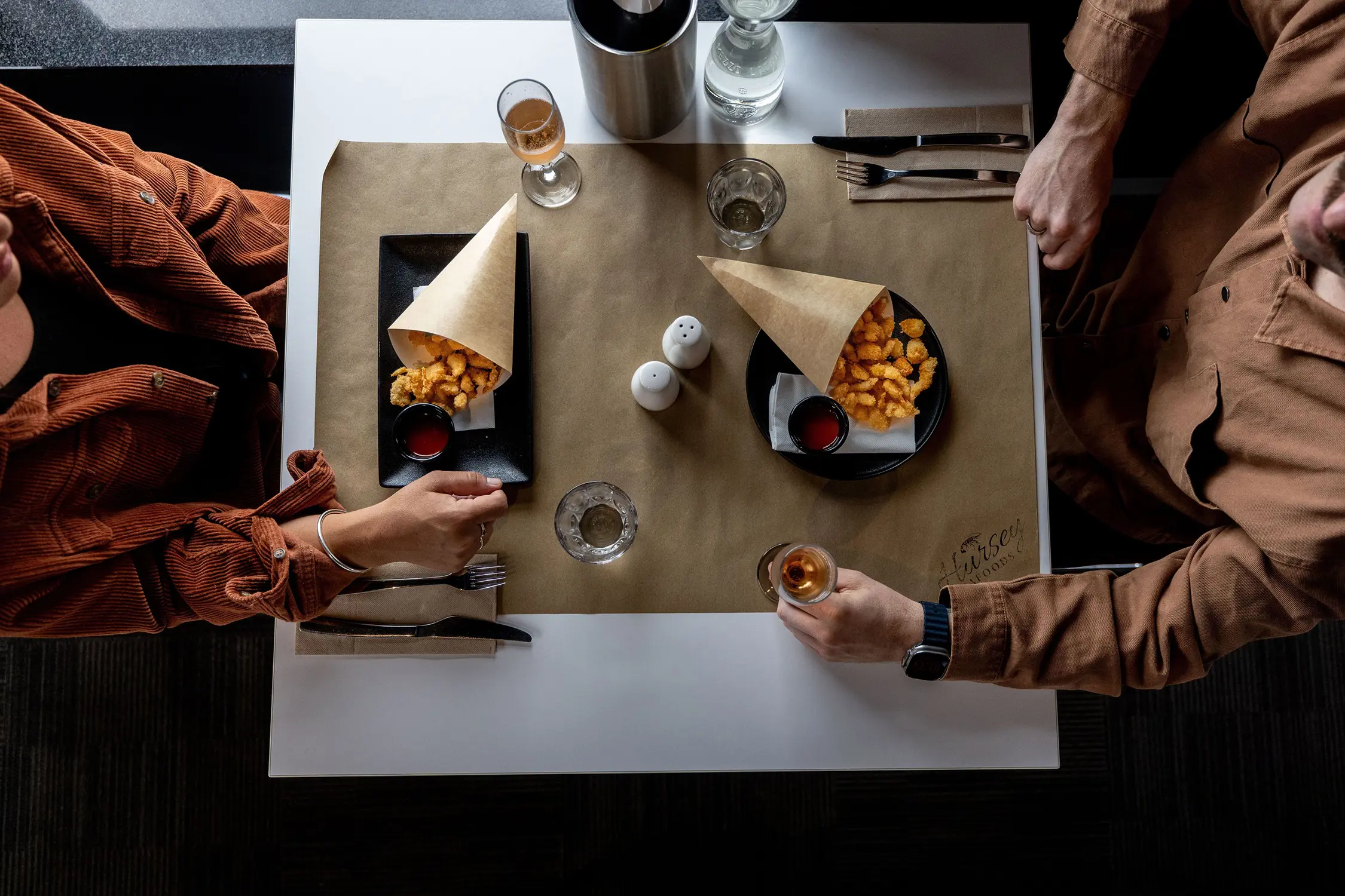 Two diners reach across a table, covered with butcher's paper, to paper cones of tasty fried seafood in front of them.