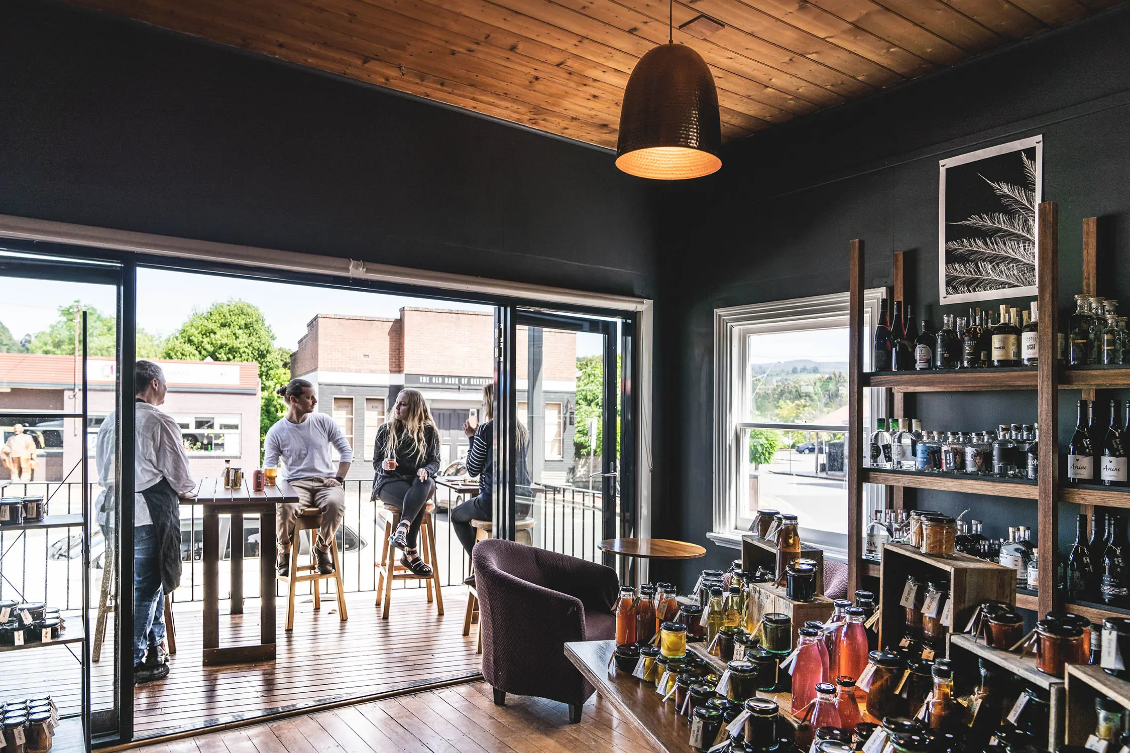 The interior shows a dining and providore space with pendant lighting, open doors and a well-stocked wall shelf filled with jars, preserves and drinks.