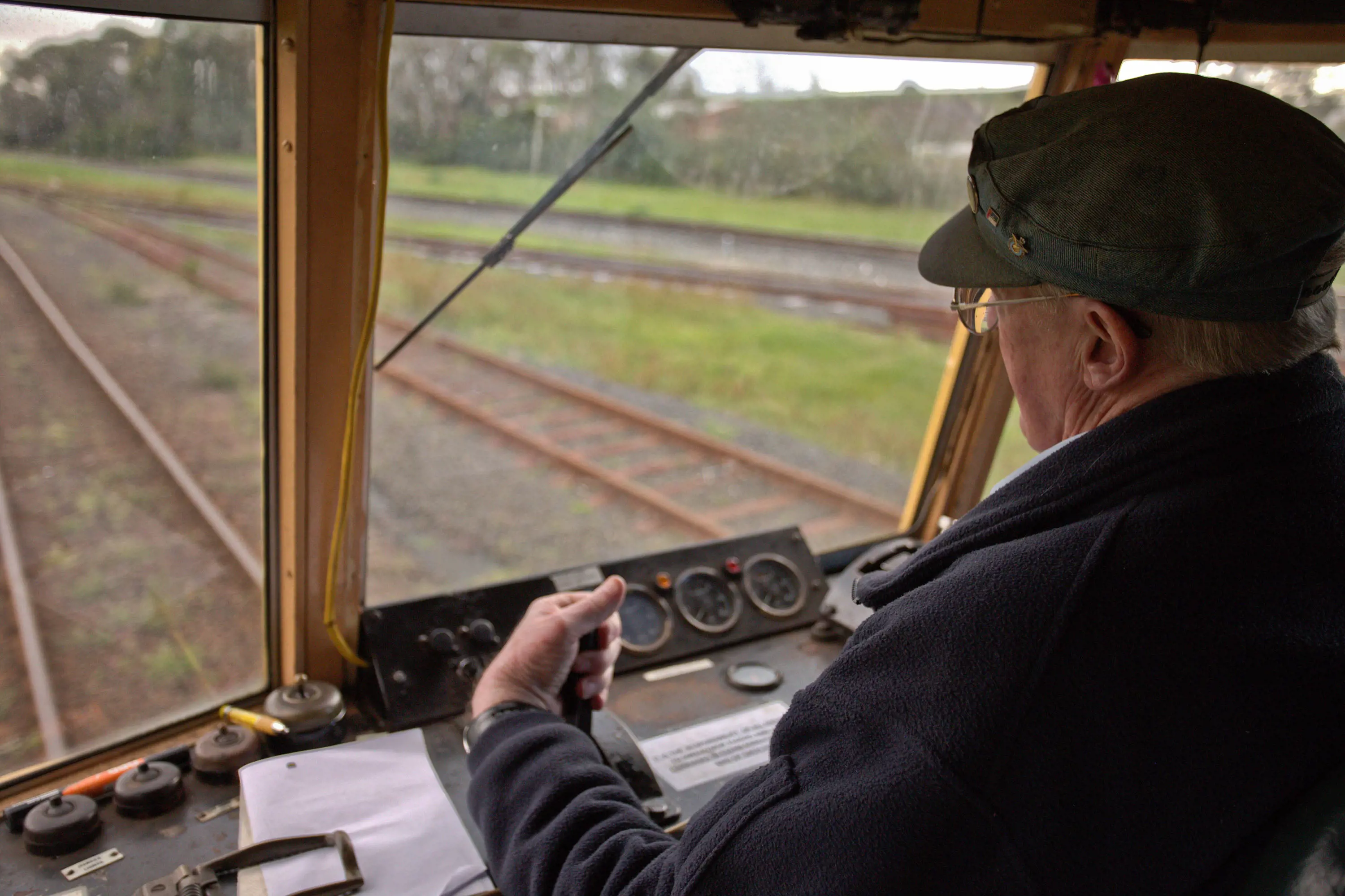 Looking over the shoulder of a man in the driver's cab of a vintage-looking train, with lots of buttons and dials. The track is laid out ahead of him through the window.