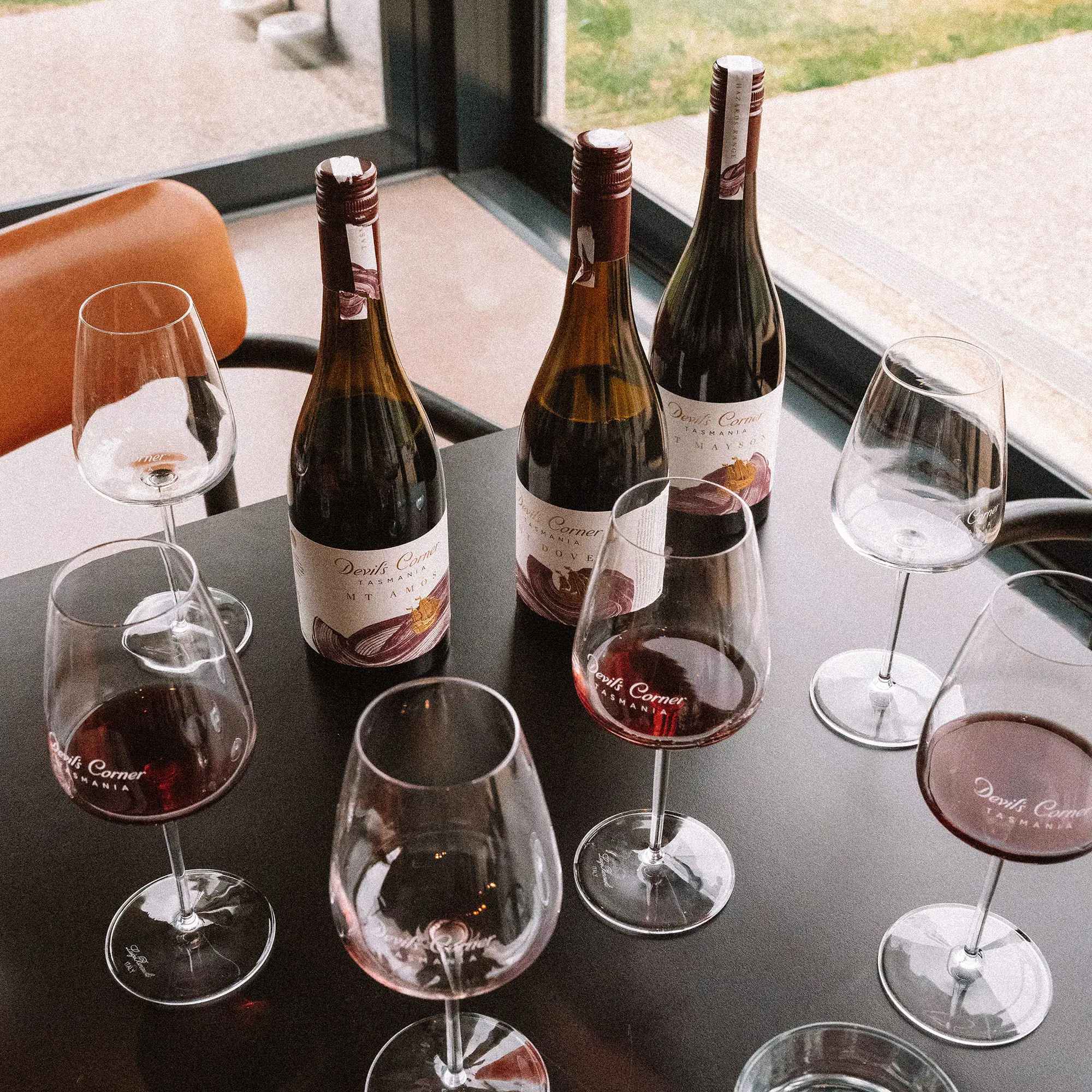 A wine tasting setup shows three dark bottles and six glasses containing red wine, on a table beside a window.