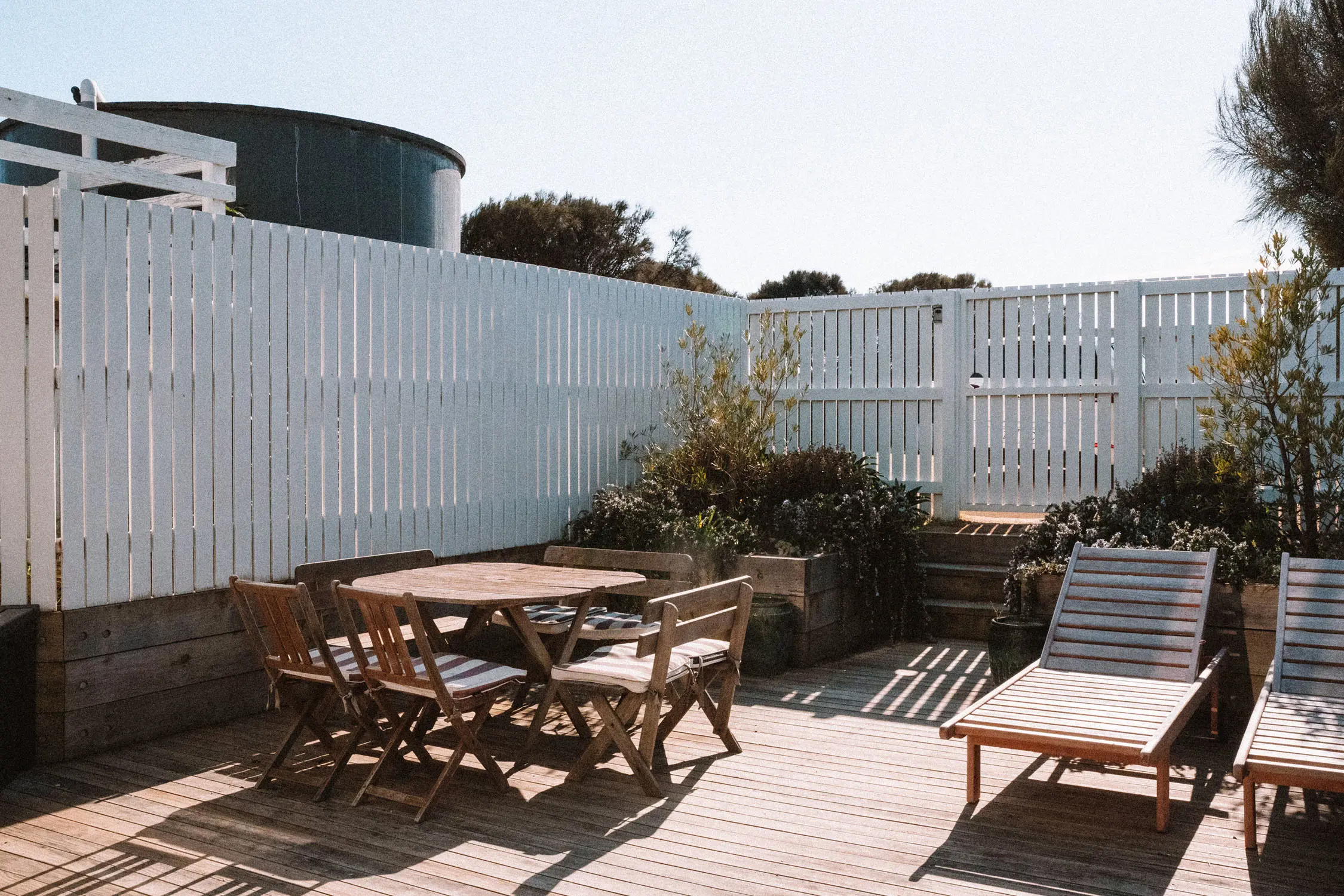An outdoor courtyard features wooden dining furniture and sun loungers on timber deck, surrounded by white vertical fencing and native plantings.