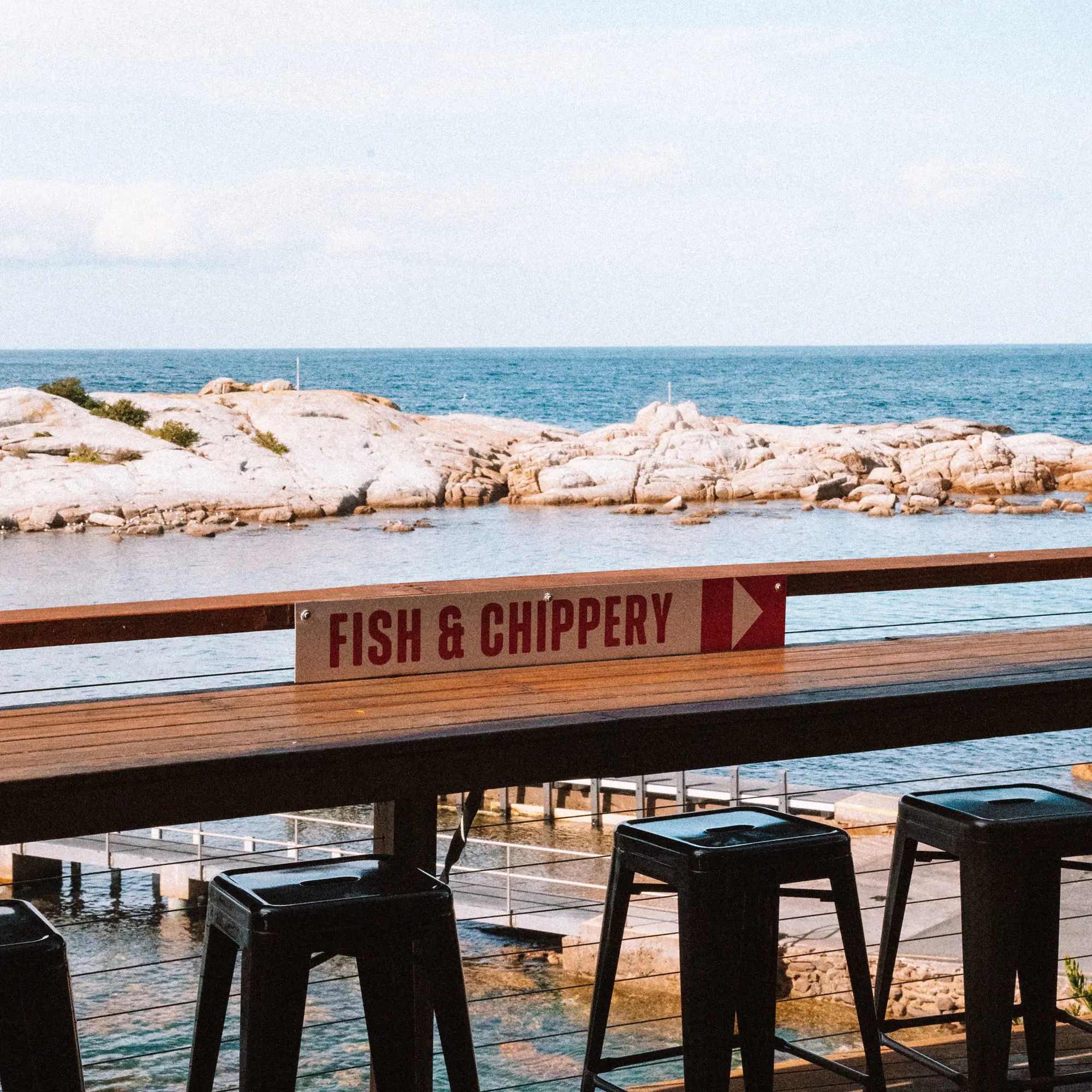 Outdoor seating features dark metal bar stools at a timber railing with a weathered sign pointing to "Fish & Chippery". Granite rocks and calm water extends to the horizon.