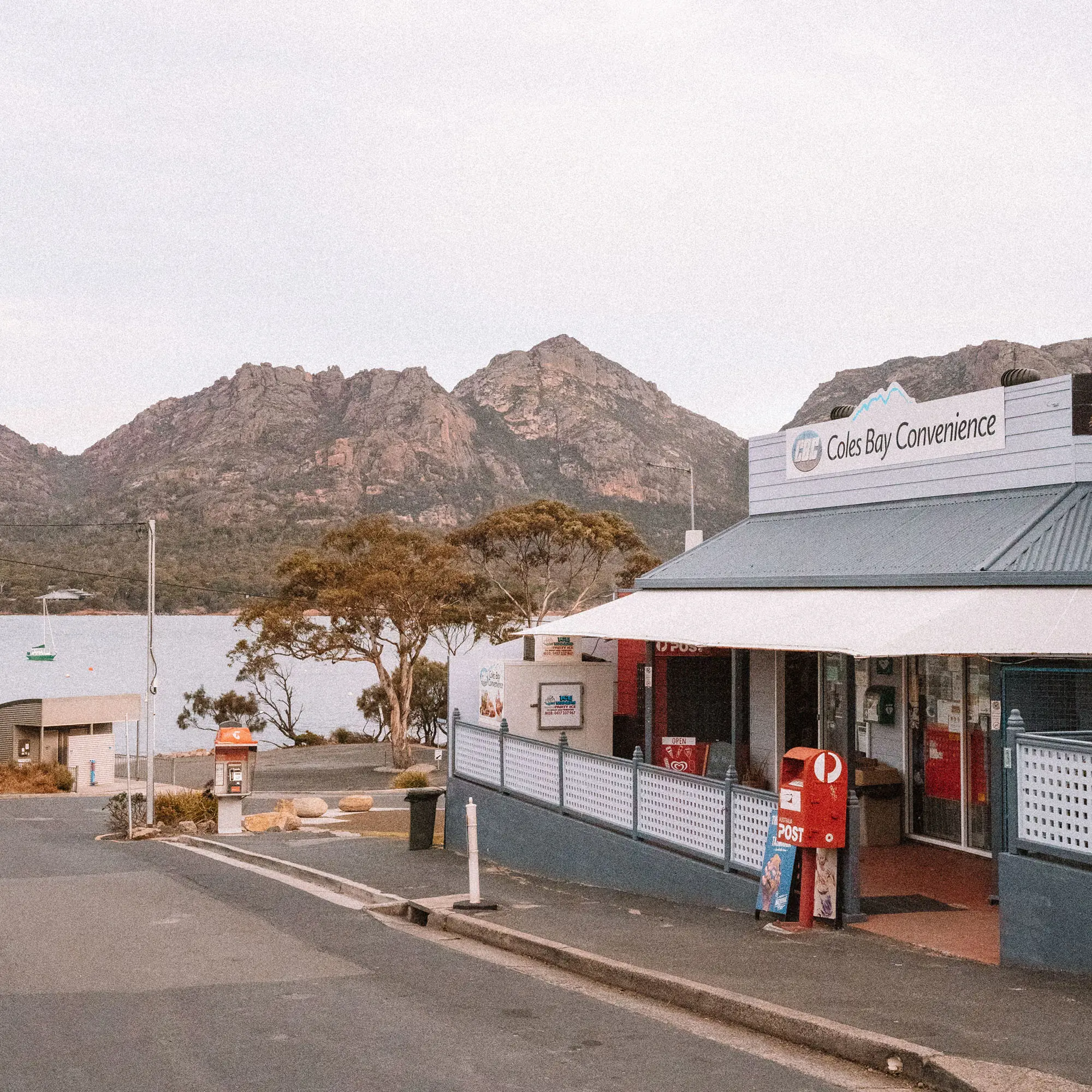 A white weatherboard shop with a metal roof, "Coles Bay Convenience", sits beside a road with mountains visible behind under overcast skies.