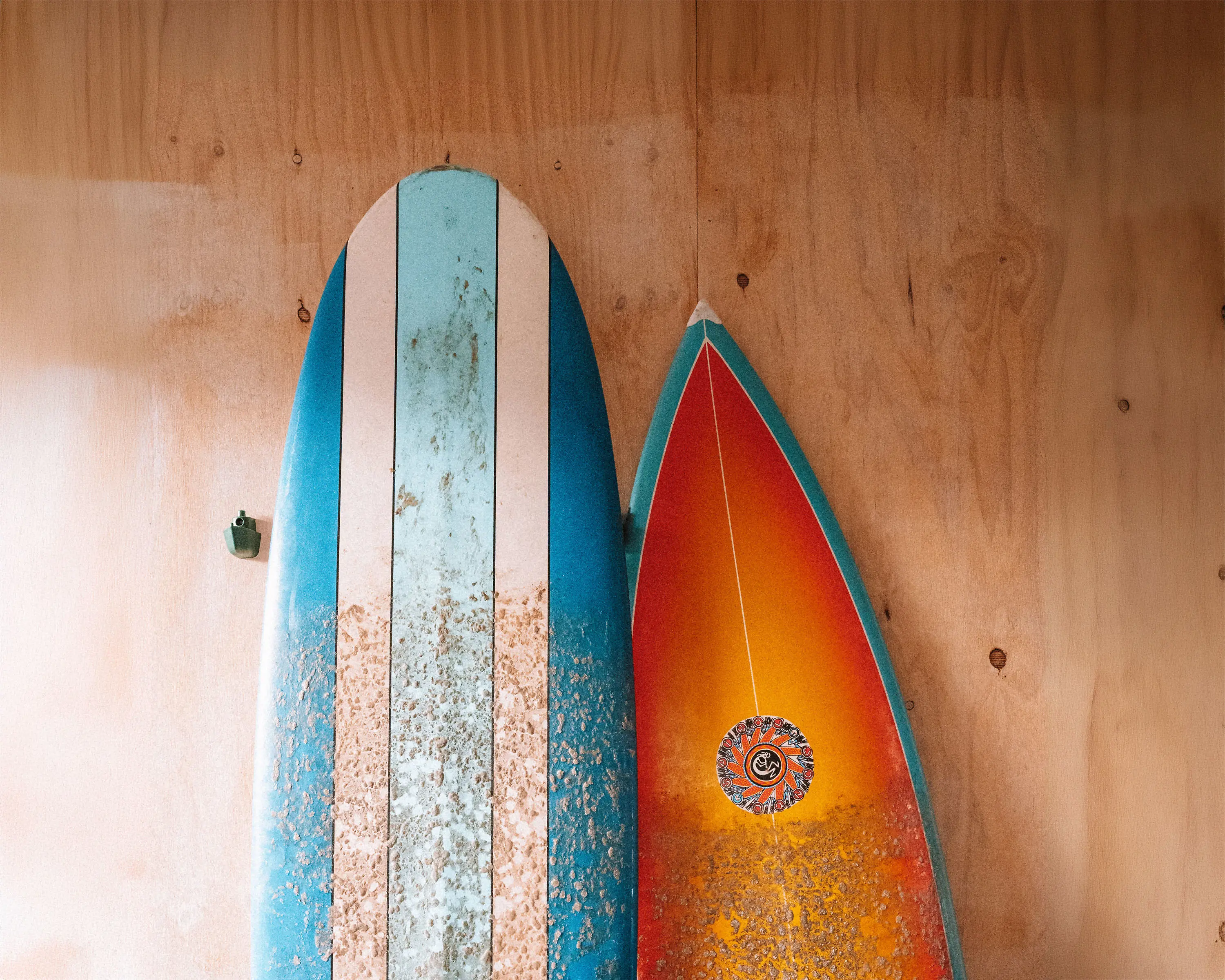 Two worn surfboards, colourful and dotted with wax, lean up against a light wooden-clad wall.