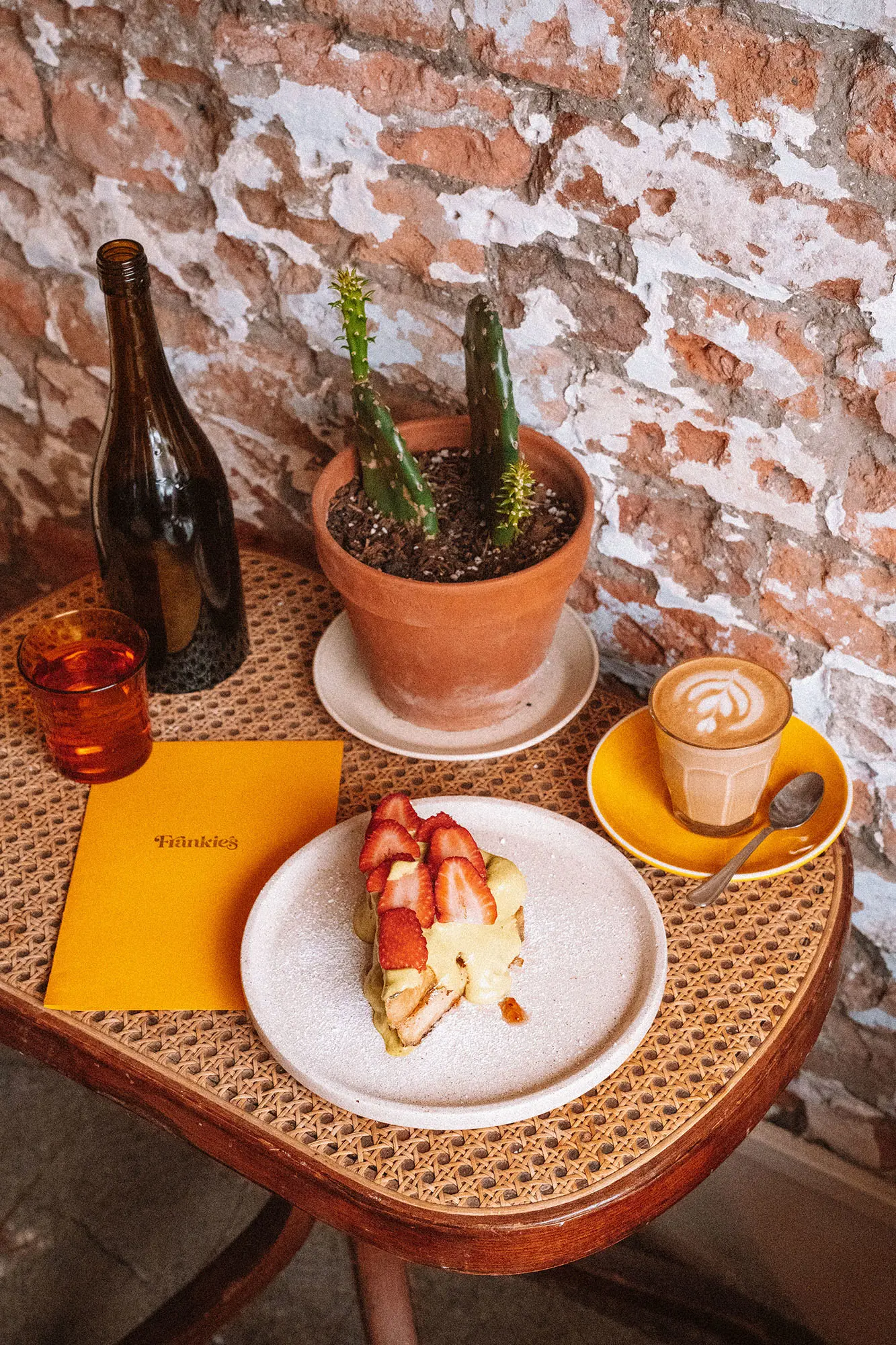 A small wicker table against a rustic brick wall, holding a cactus pot plant, a plate of food, a latte and an orange menu with 'Frankie's' on the front.