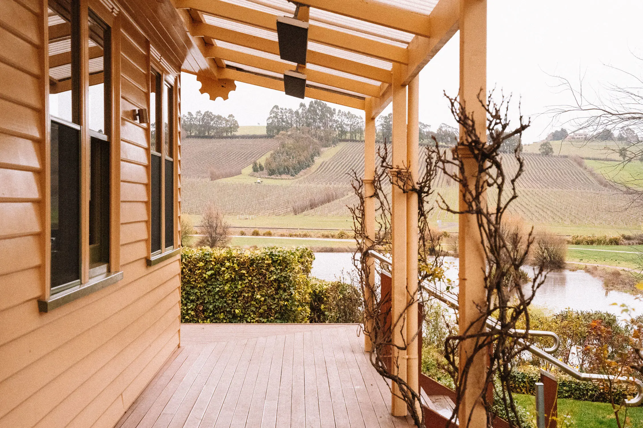 Looking down the wrap-around patio of a yellow weatherboard building, over sprawling hills of vineyards and a small lake.