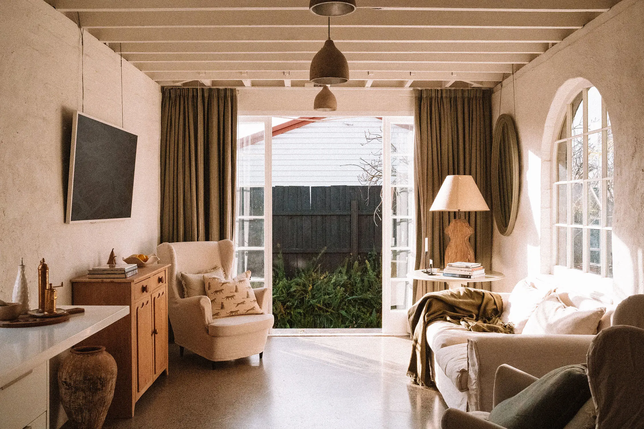 The interior of a loungeroom, with textured walls, architectural details like an arched window, and comfy olive green and cream furnishings. The French double doors are open to the garden.