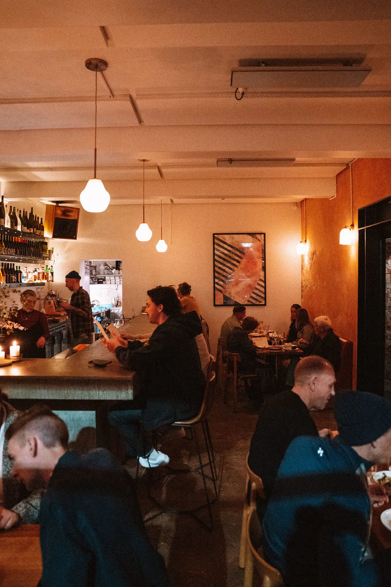 The interior of a cosy bar, busy with full tables and those seated at the counter ready to order. Low-hanging ceiling pendants cast a warm glow.