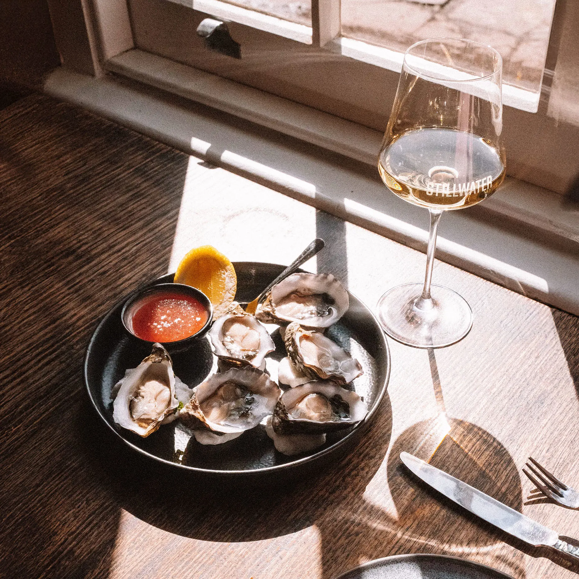 A plate of oysters and a glass of wine sit on a wooden table by a sash window, with daylight streaming in over the scene.