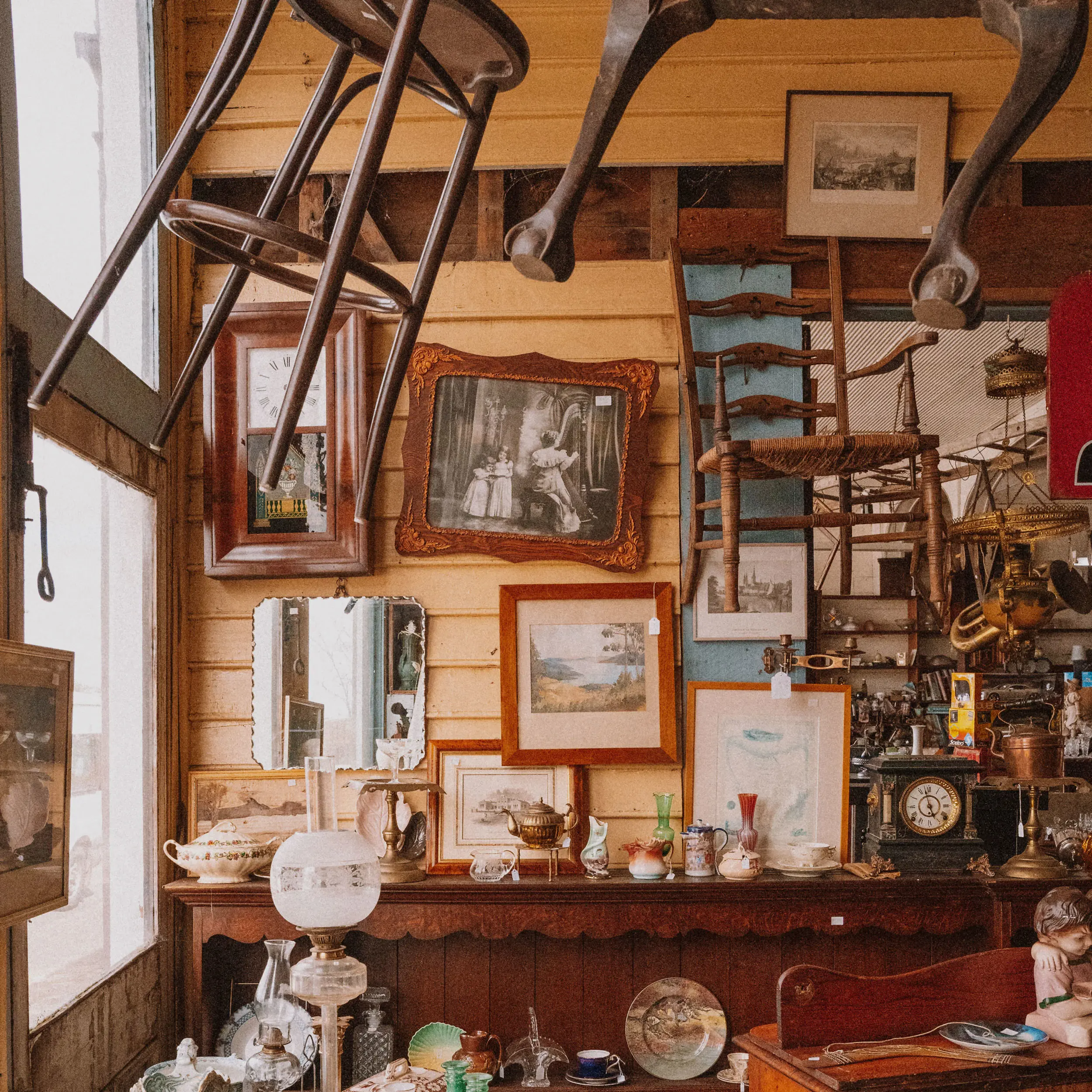 The interior of an antiques shop. The weatherboard wall is covered in hanging pictures and mirrors, chairs are suspended at angles from the ceiling, and a wooden sideboard is covered in treasures.