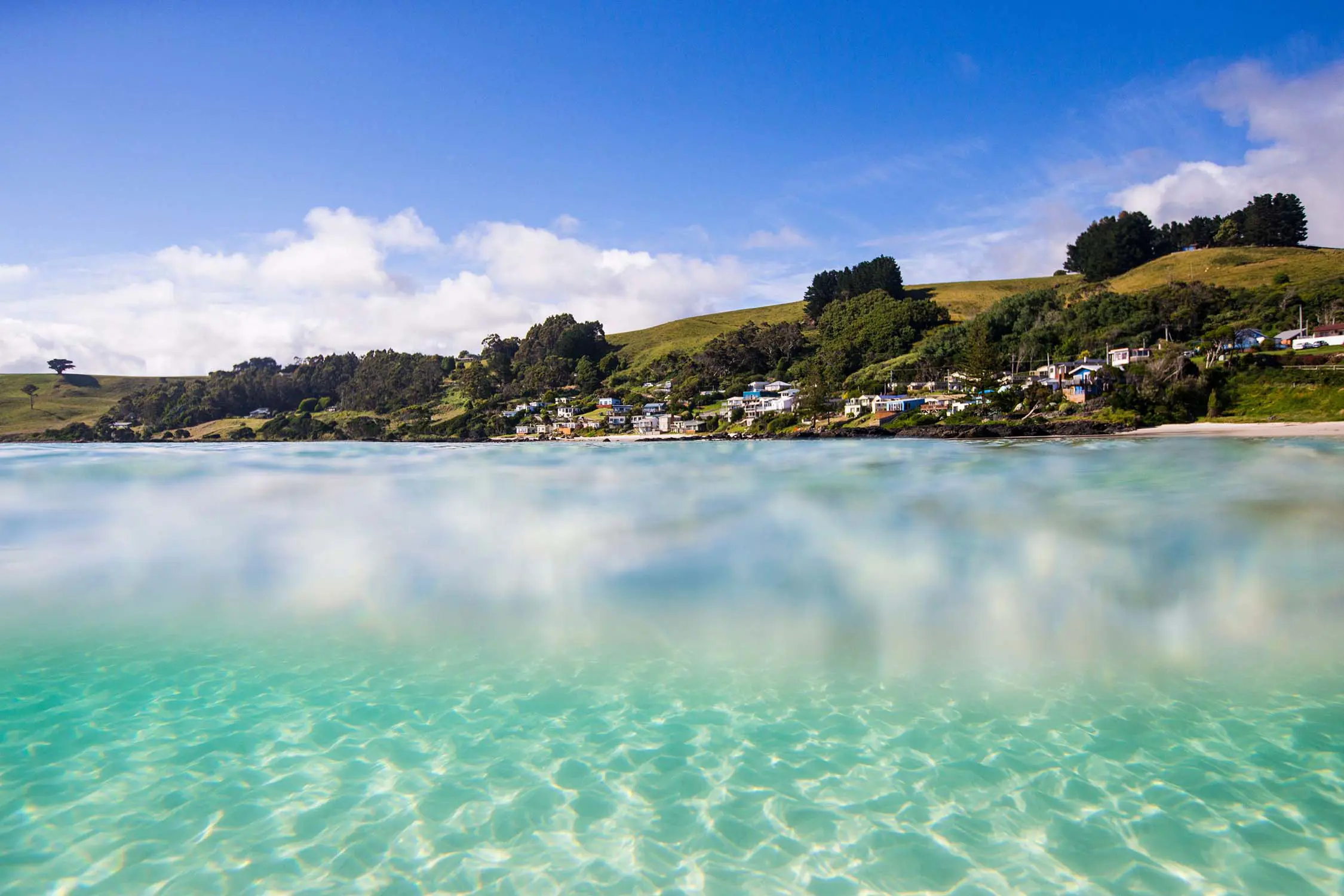 An image taken half-submerged in the crystal-clear waters of a beach on a sunny day. Above the waterline, a small town sits on some hills, and below, the water's ripples reflect onto the sandy seabed.