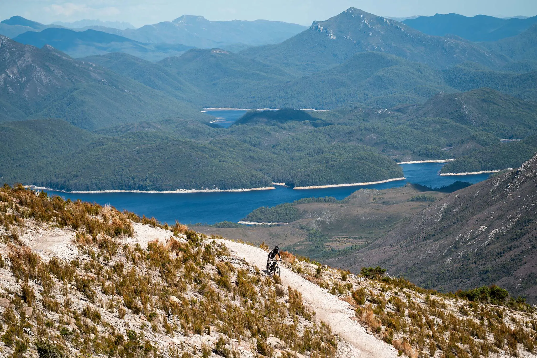 A person on a mountain bike rides down a sandy dirt path along the side of a hill, with a stunning mountain backdrop.
