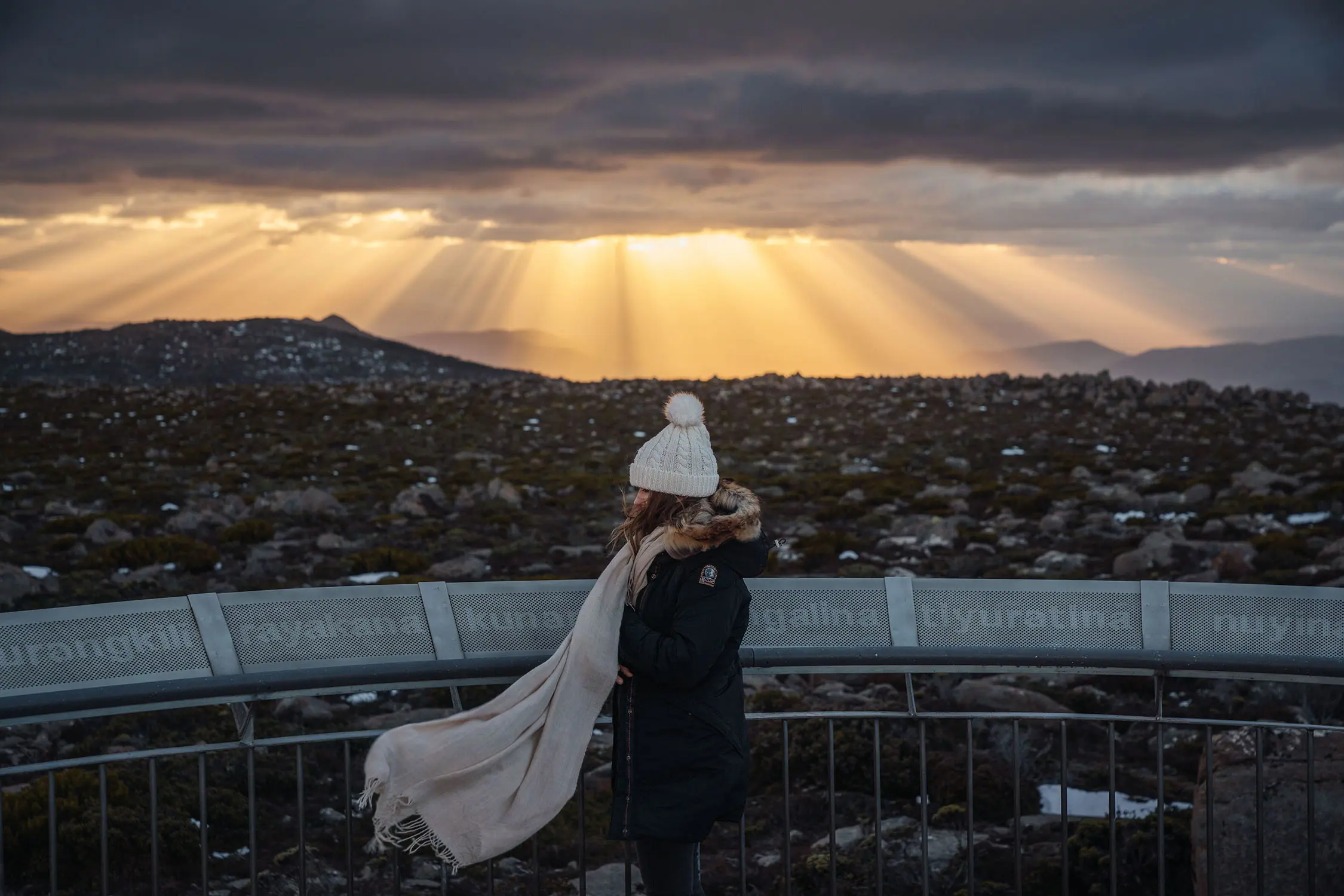 A woman in a beanie and scarf, her scarf blowing around her in the wind, standing on a mountain lookout overlooking a city. The sun shines a few rays through the heavy cloud above.