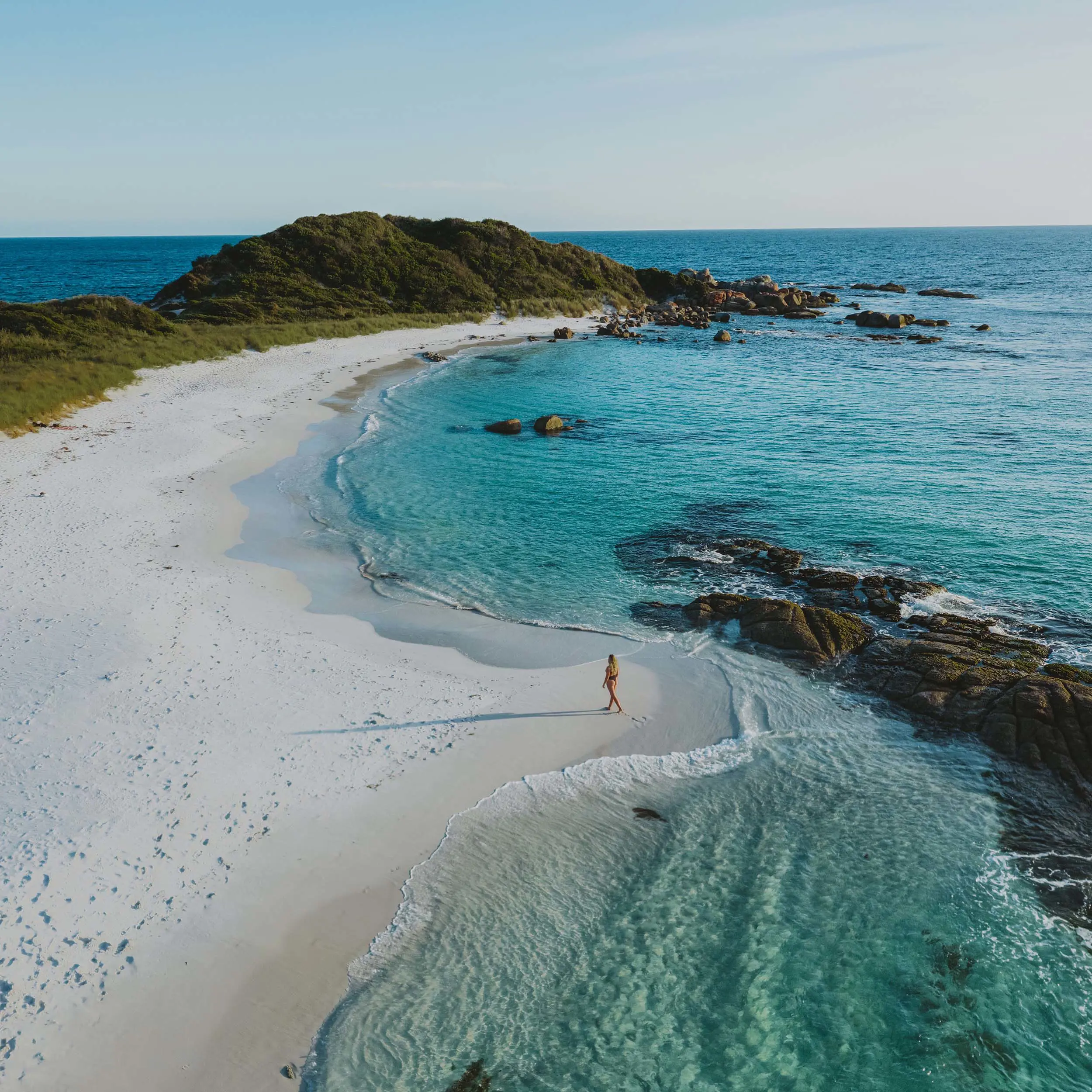 A woman walks along a picturesque curving white beach. The water is crystal clear blue, dotted with rocks and a forested hill rising at the end of the spit of land.
