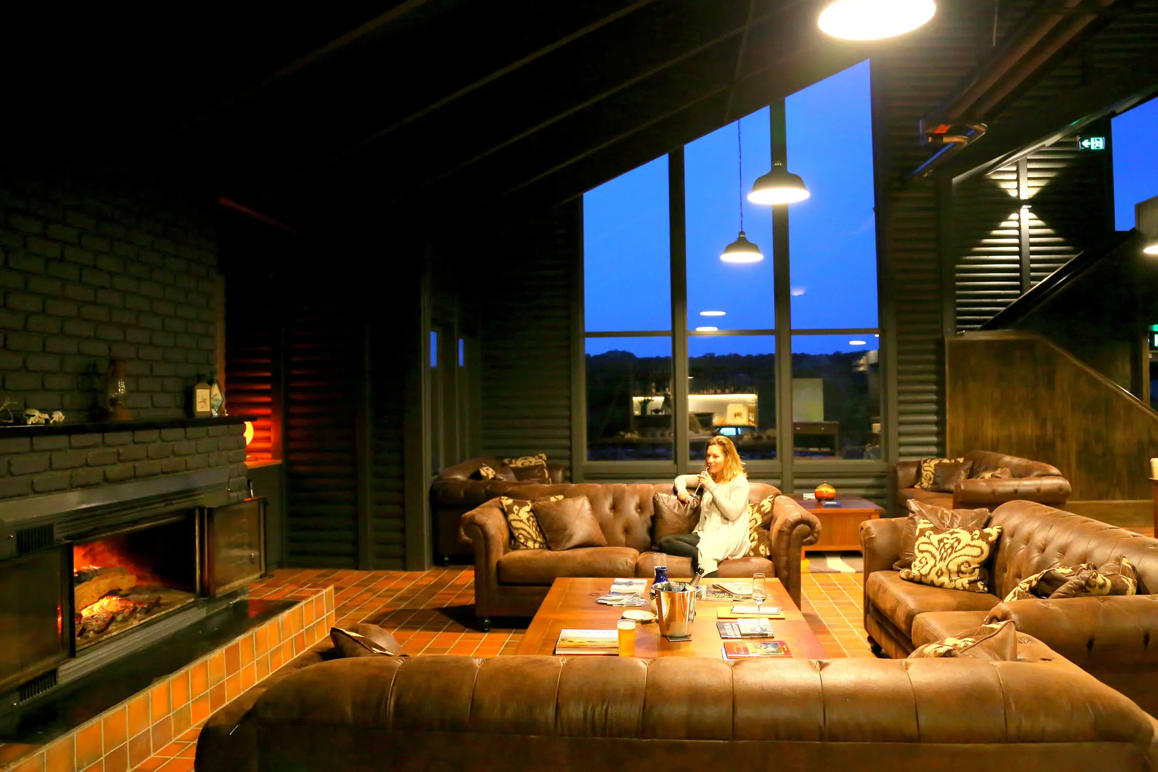 In an expansive lounge room with three brown leather chesterfield couches, a woman is relaxing in front of a crackling fireplace.