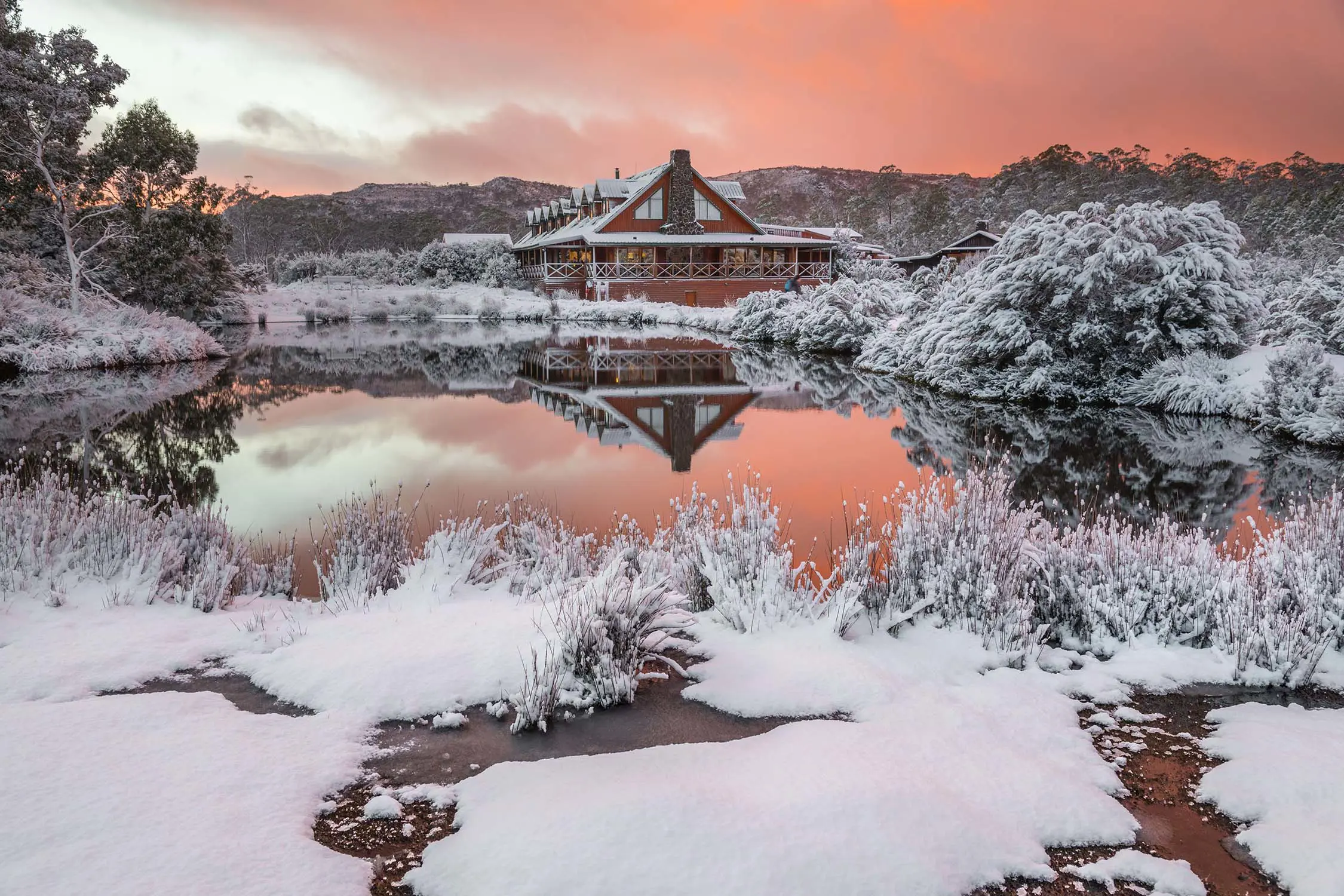 A snow-swept landscape with an alpine lodge in the background. The water is mirror-still, reflecting the scene and the orange sunset sky above.