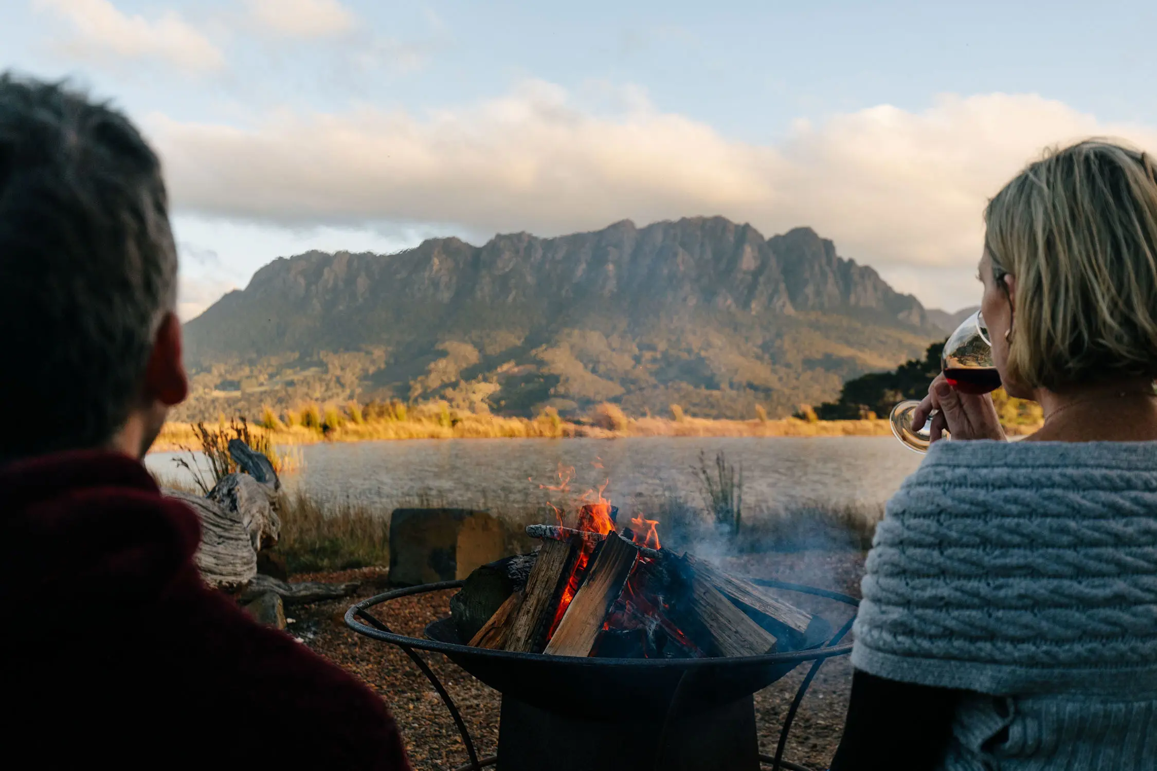 Two people sipping wine are facing away from the camera, over an outdoor firepit. A mountain range is lit by sun over the water in the background.