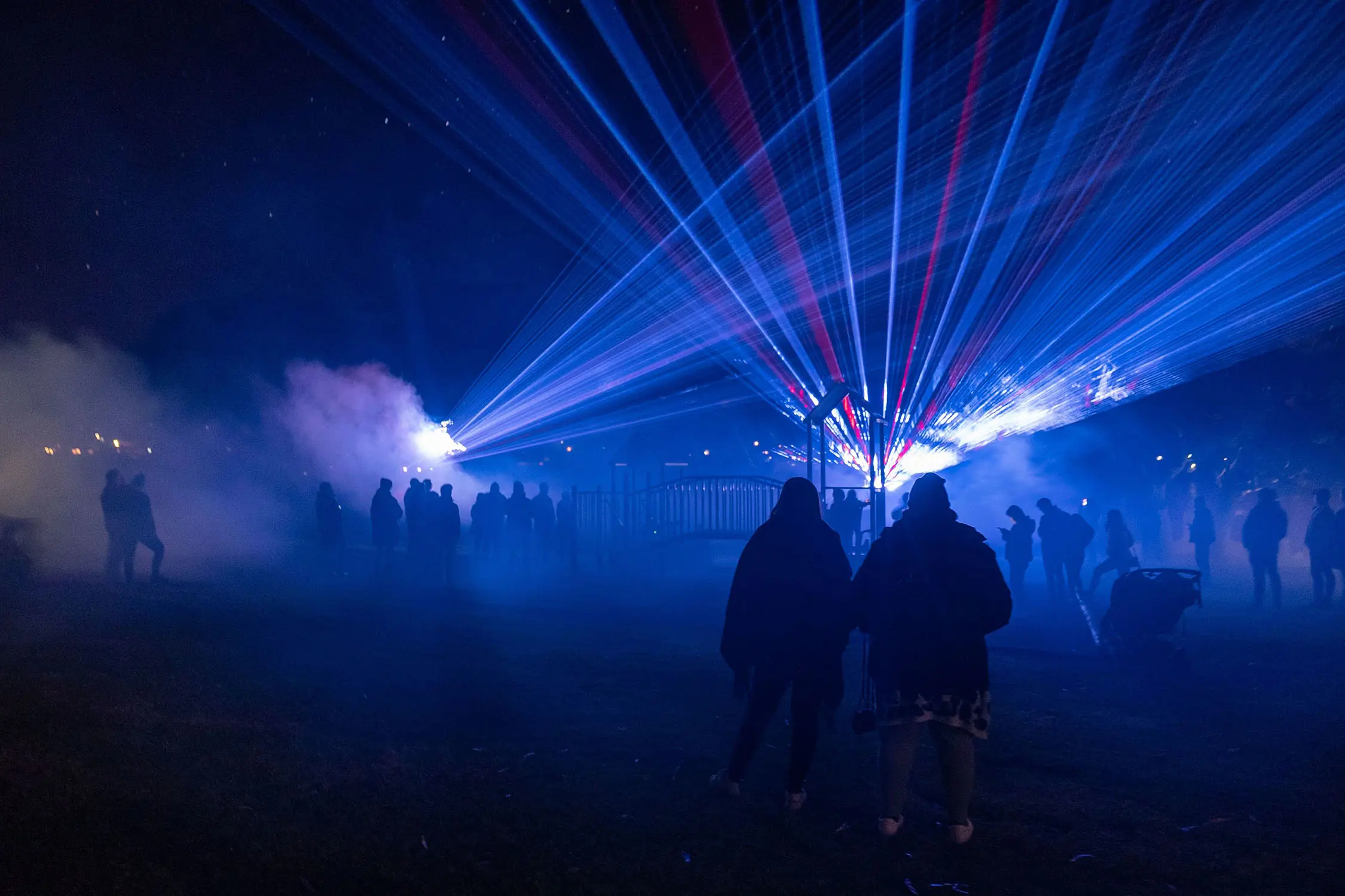 A couple stand in a park lit mysteriously by the blue and red laser lights that are cast over their heads. Many more people mill around, shrouded by smoke.
