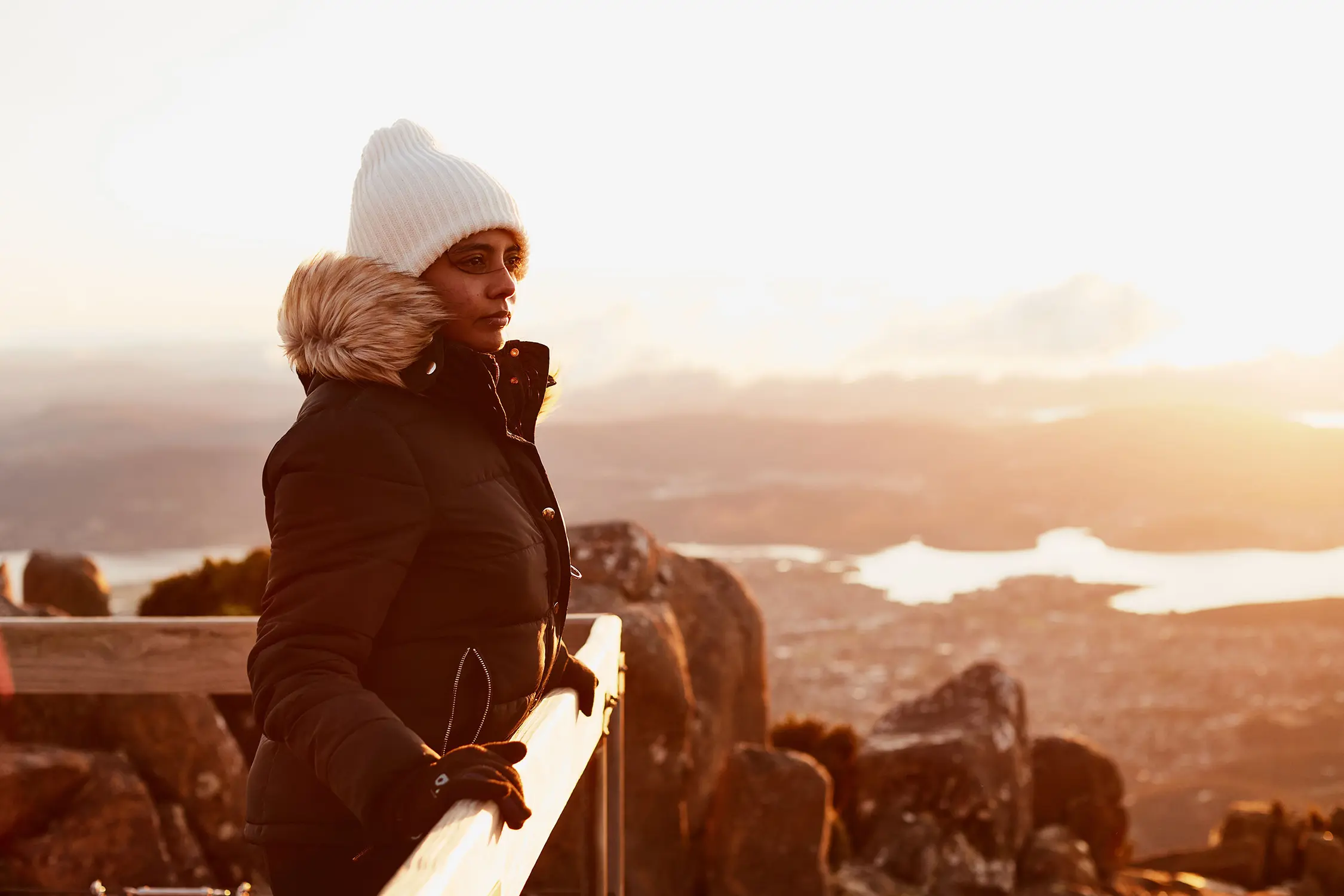 A woman in a beanie, gloves and thick winter coat stands on a mountain viewing platform, looking out over the city and river below.