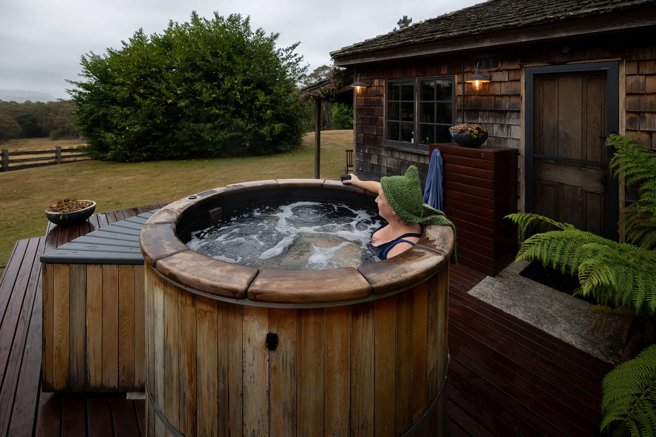 A person in a beanie and bathers, relaxing in a warm wooden hot tub outdoors, next to a small wooden cottage.. 