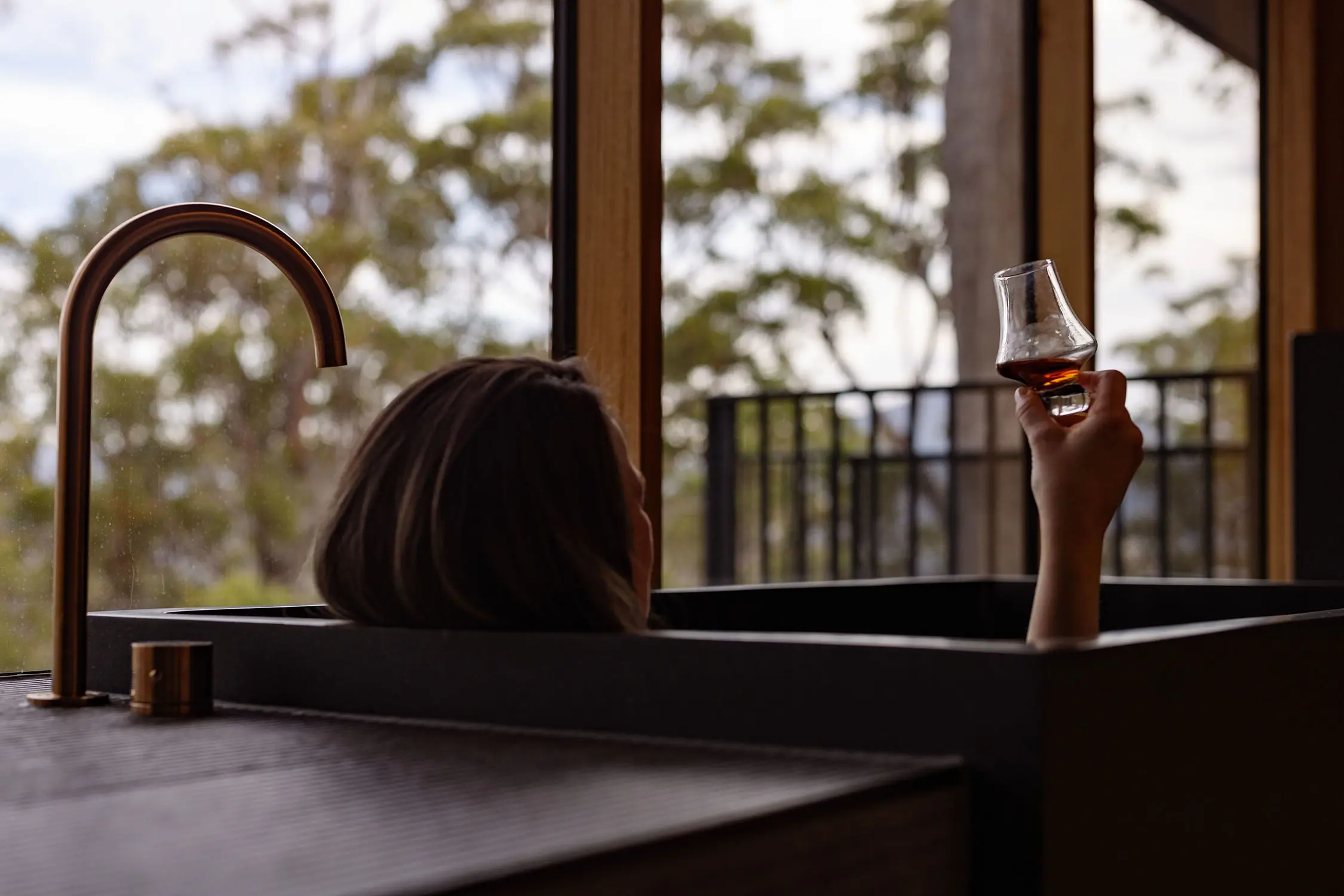 A person reclines in a bathtub near a large window, holding a glass of wine raised in the air. Outside, trees fill the serene view.
