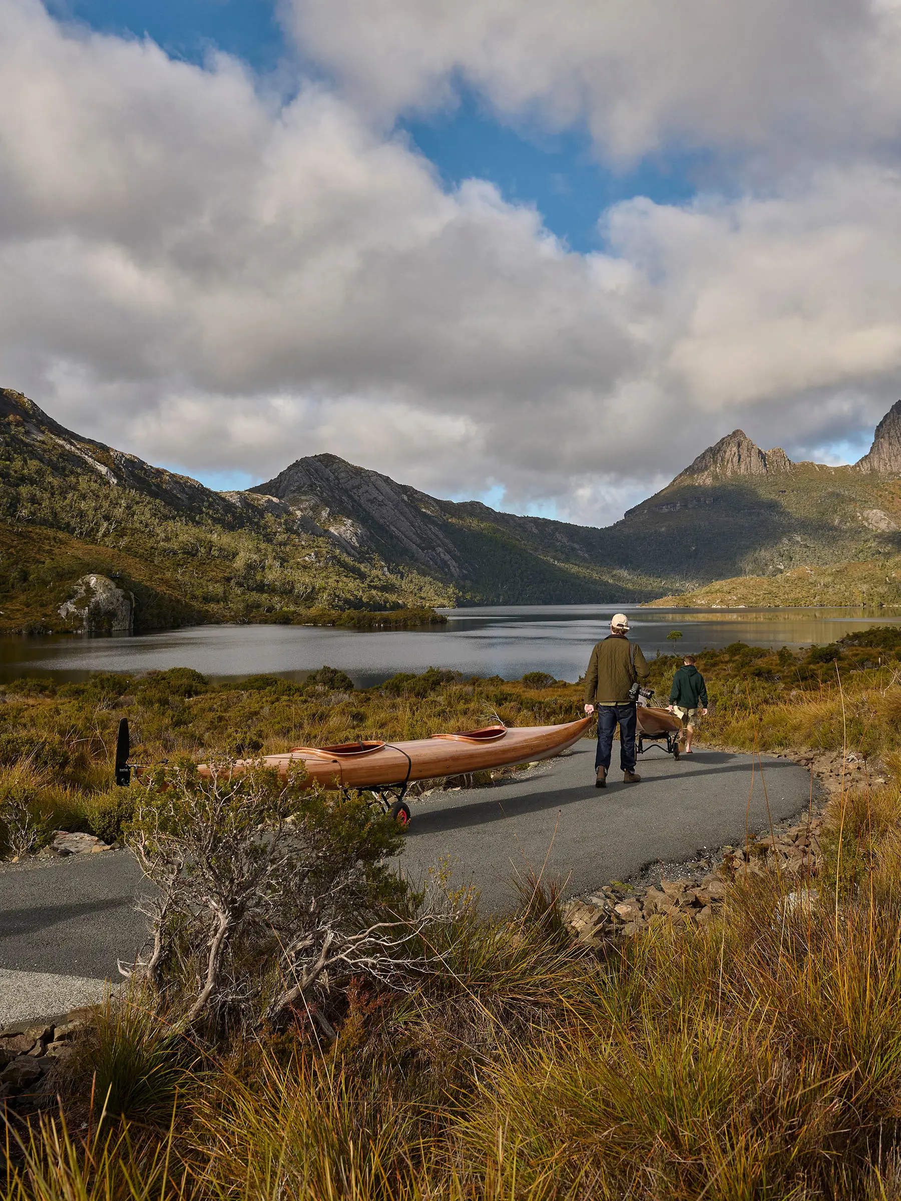 People stand on a mountain road, hauling wooden kayaks, looking towards a glassy lake with dramatic mountain peaks visible across the water.