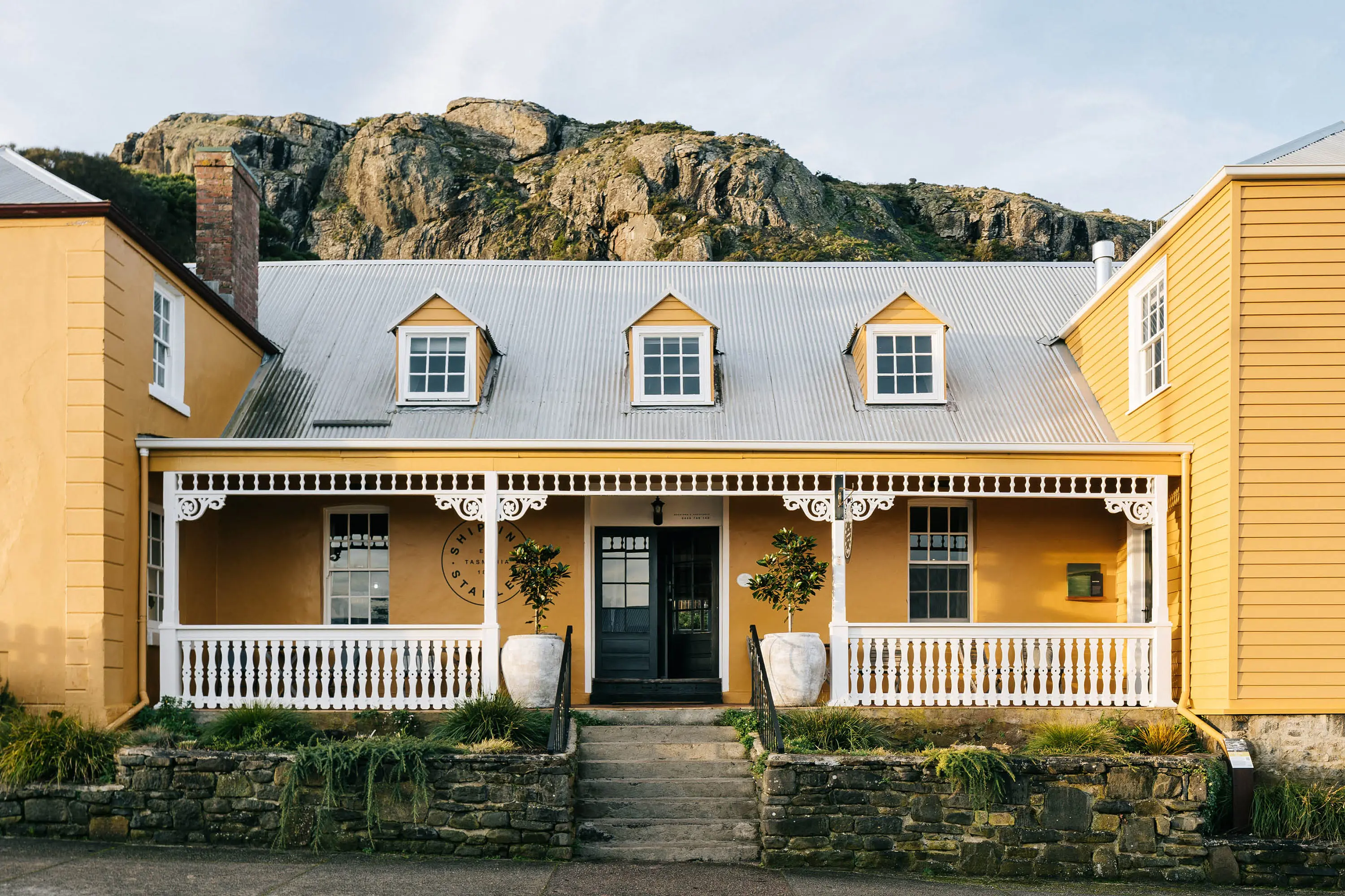 A century-old colonial-style building with yellow walls and white trim features a white picket fence, with bush-covered mountains visible behind.
