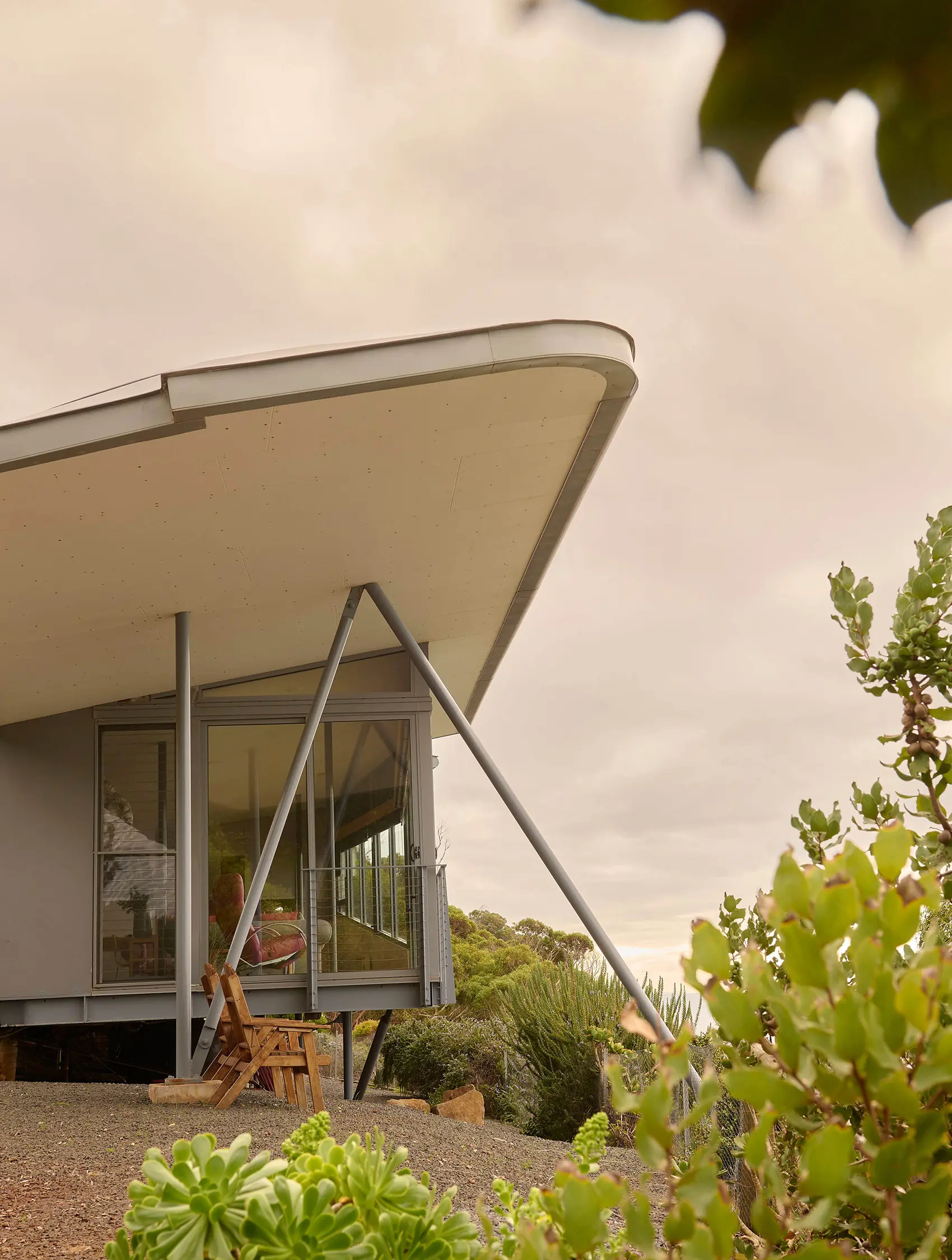 A modern angular building, with a soaring cantilevered overhanging roof and floor-to-ceiling windows, sits among native vegetation on a hillside overlooking the landscape.