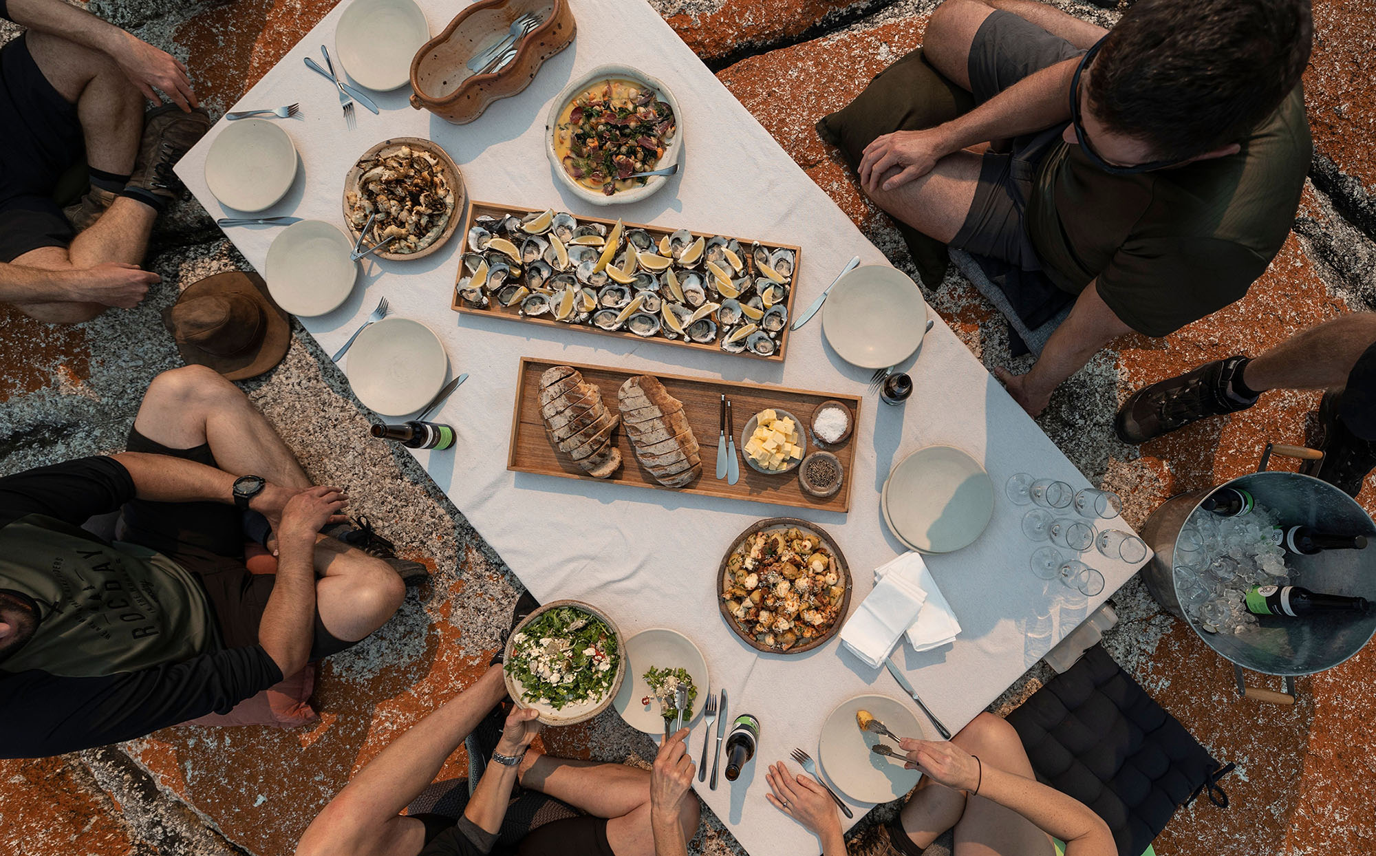 A top-down view of a group around a picnic table perched on an orange-tinged rock. The table is full of oysters, fish and other plates and drinks.