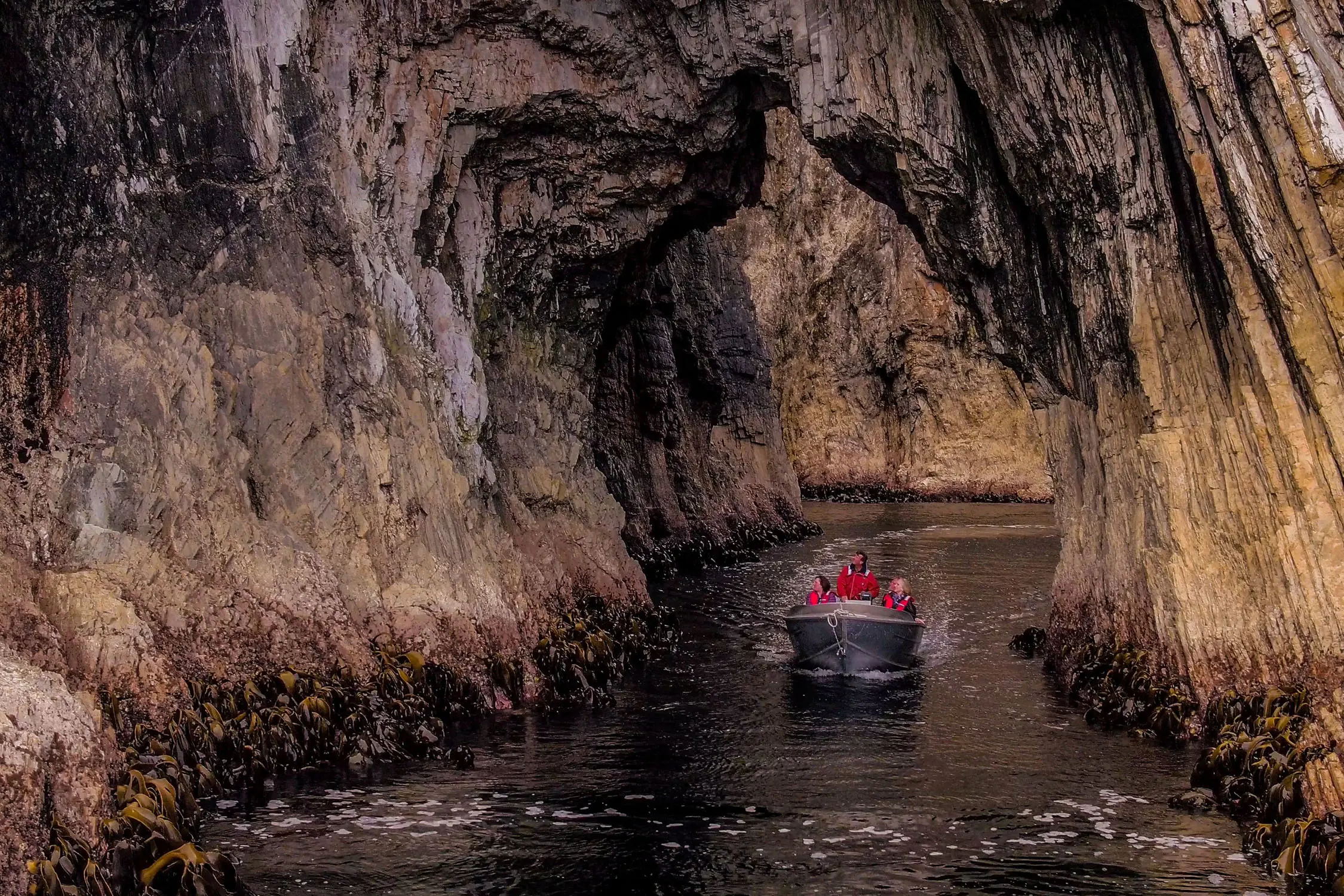 A small group in red jackets ride a small silver boat through a stunning natural rock arch, gazing up at the shapes around them.