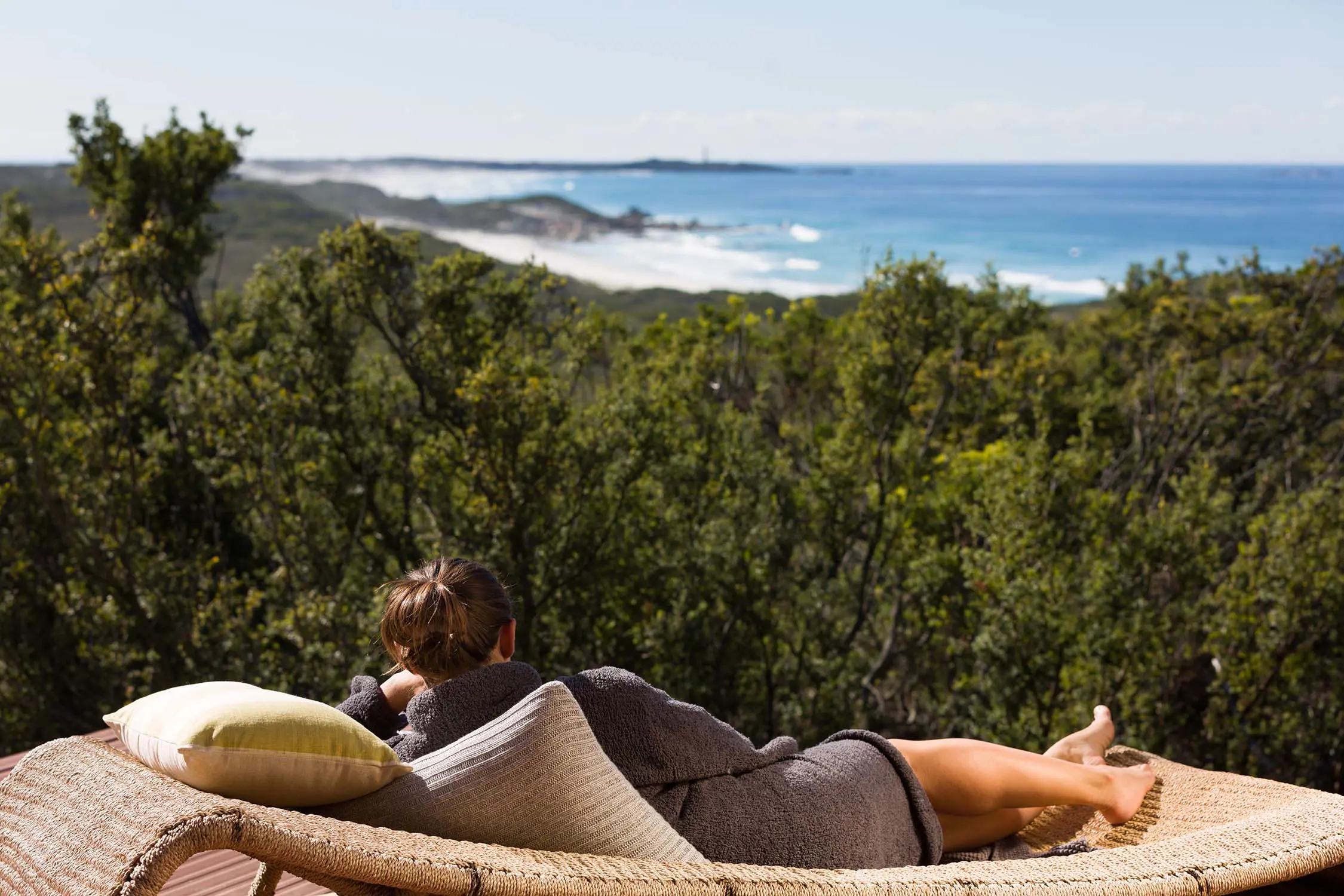 A woman reclines outdoors on a rattan sun lounger, looking over the dense bush to the blue ocean coastline beyond.