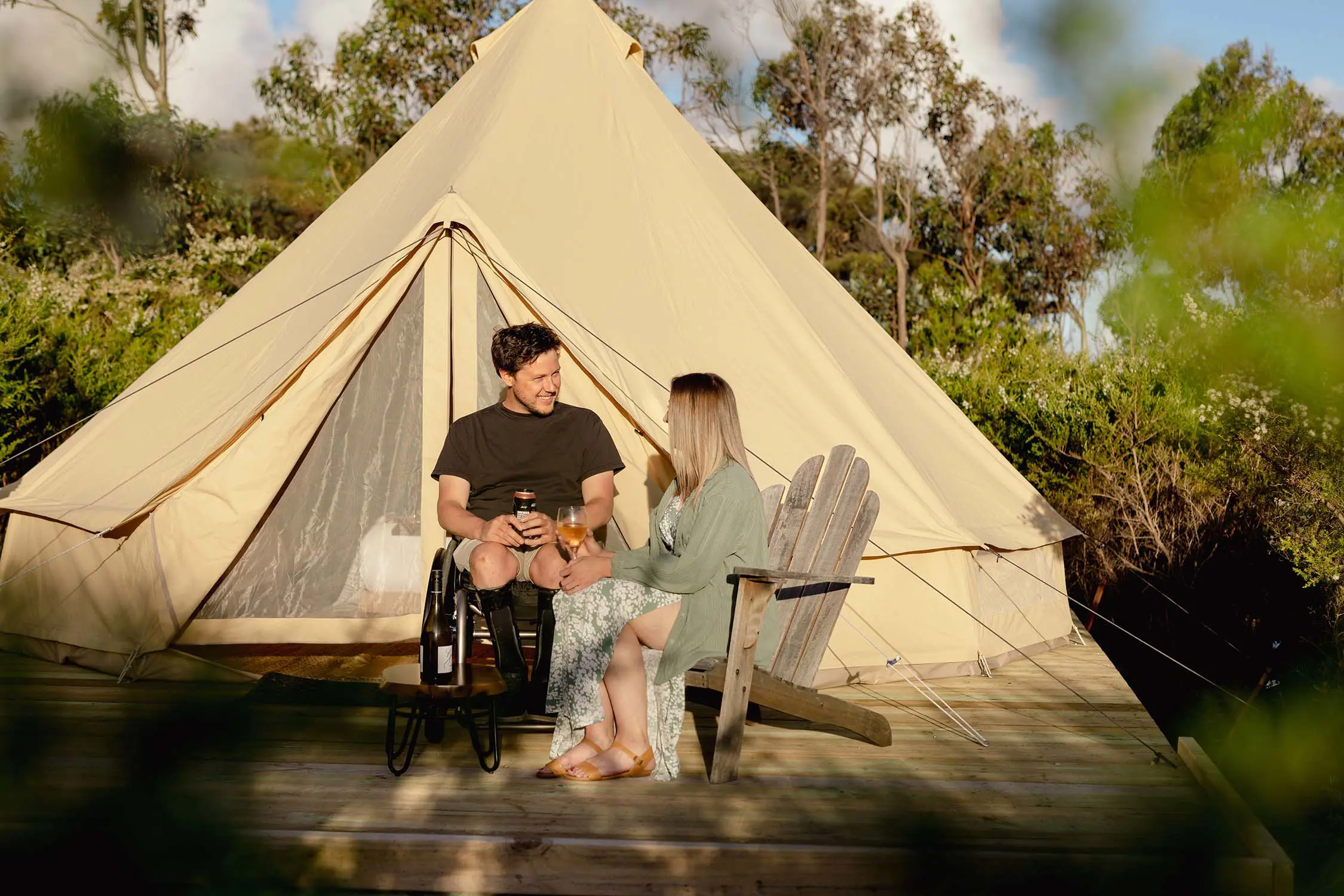 A young couple, one in a wheelchair, sit on a wooden platform in front of a large dome tent surrounded by low-level bushlands.