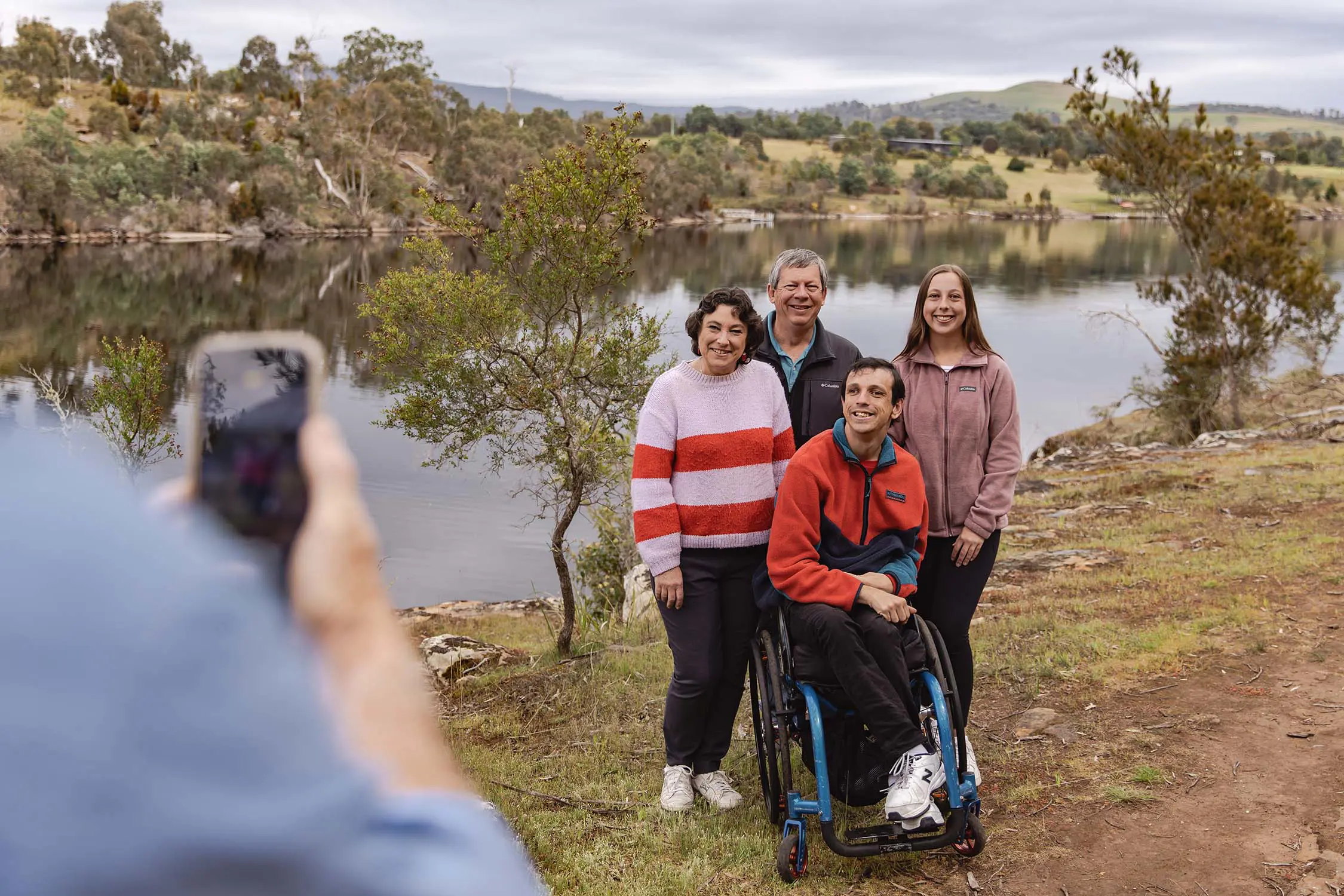 Four people pose for a photo on a small path running next to a river surrounded by trees and bush scrub. in front of a herd of sheep. One man sits in a teal blue wheelchair.