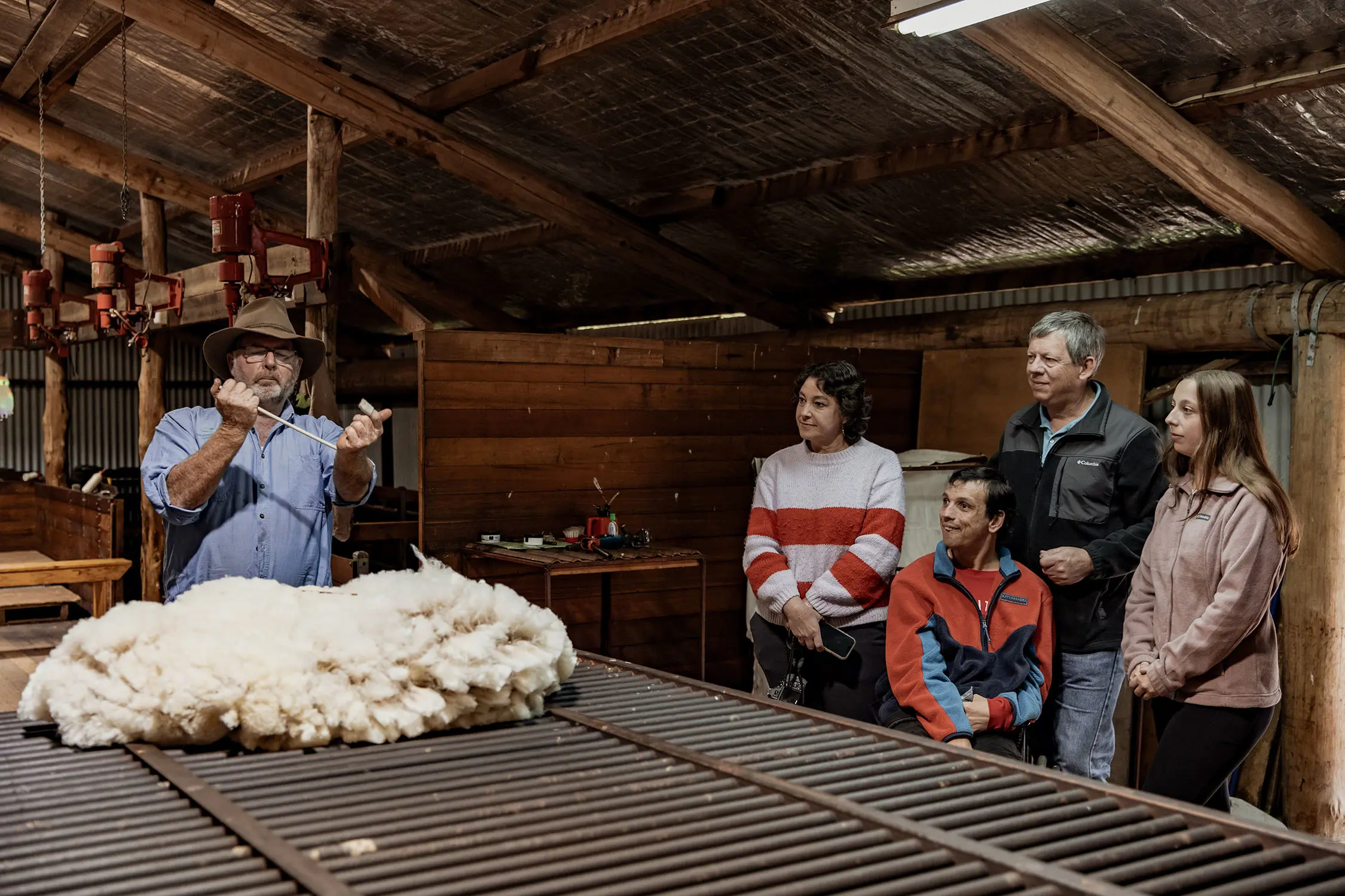 Four people stand inside a shearing shed, watching a man demonstrate with a wool fleece on the table in front of him.