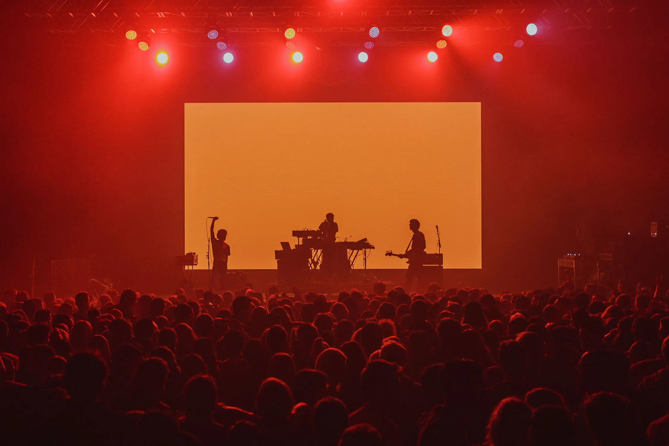 In a hazy live music venue, a crowd of silhouetted people dance to a band onstage. They are lit by red, moody lighting and framed by a bright orange screen behind them.