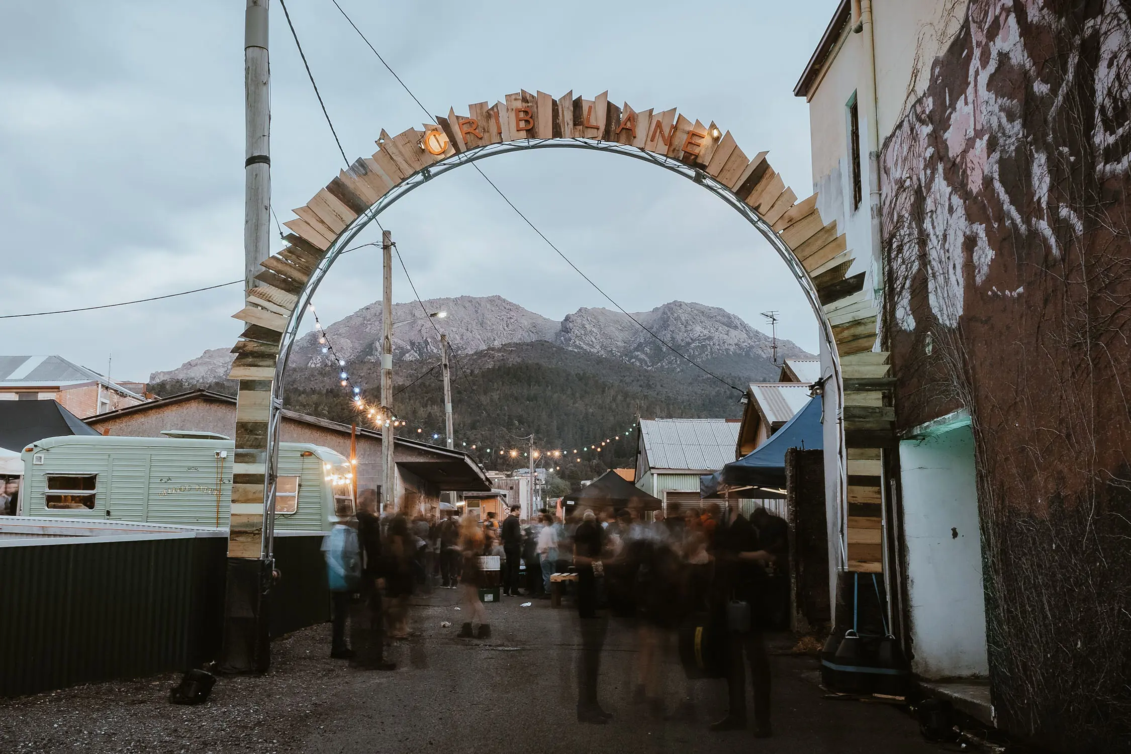 A rustic festival alleyway with caravans, tin sheds and fairy lights is framed by a jagged wooden archway with the letters "Crib Lane". Snow-capped mountains rise in the background.