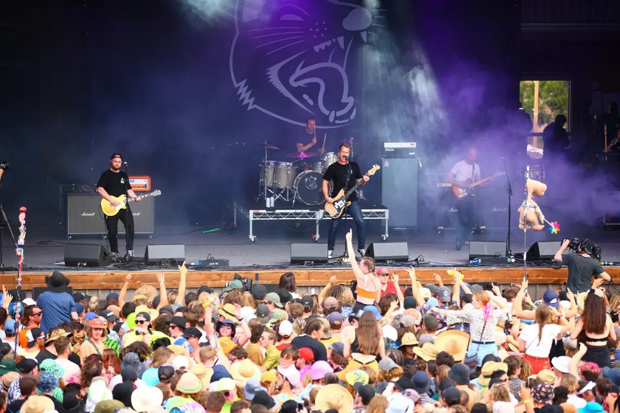 A band of four musicians plays on a festival stage. The crowd are excitedly dancing along, many with their hands in the air.