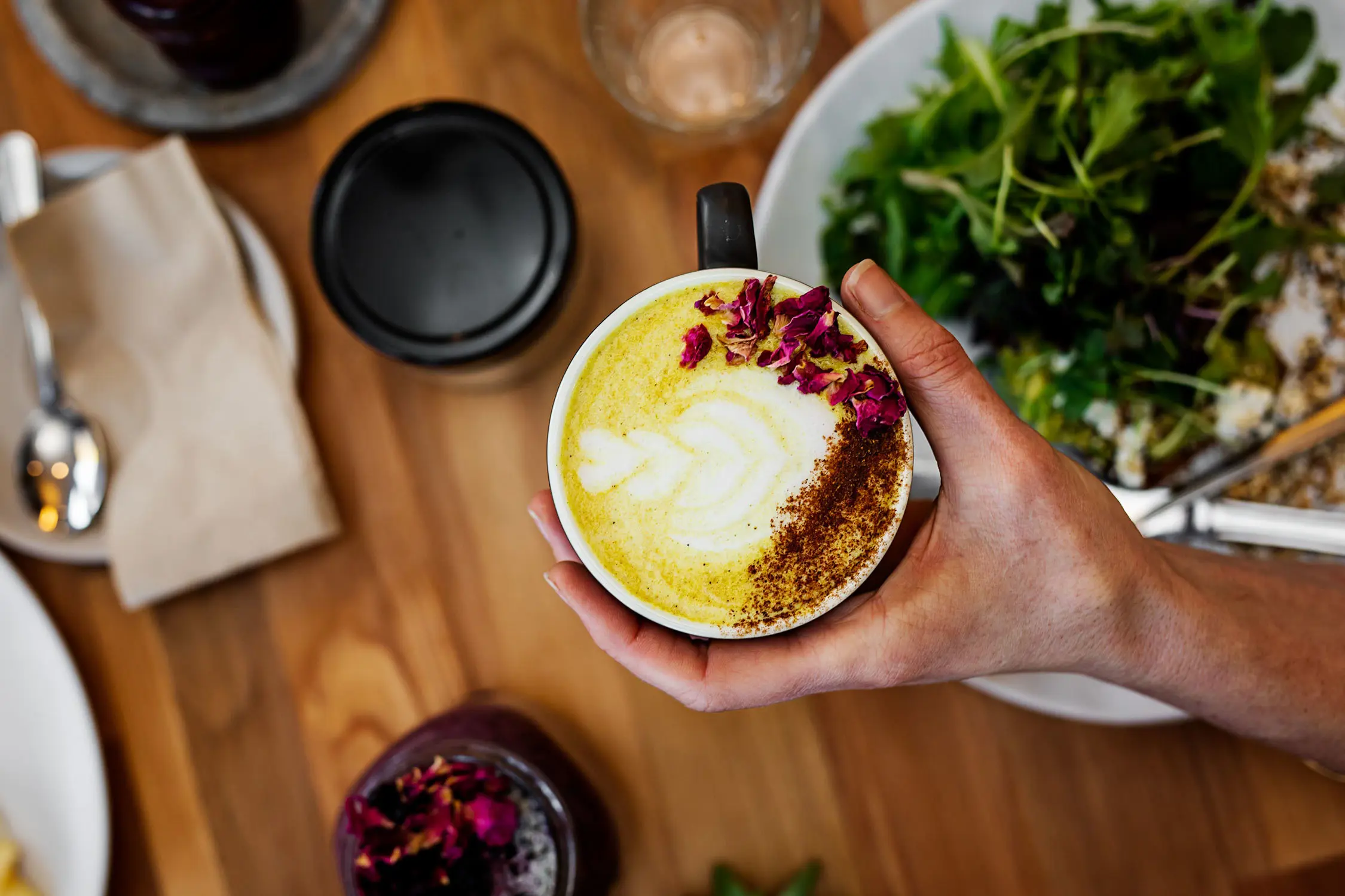 A top-down view of a person's hand holding a turmeric latte, decorated with petals and chocolate powder.