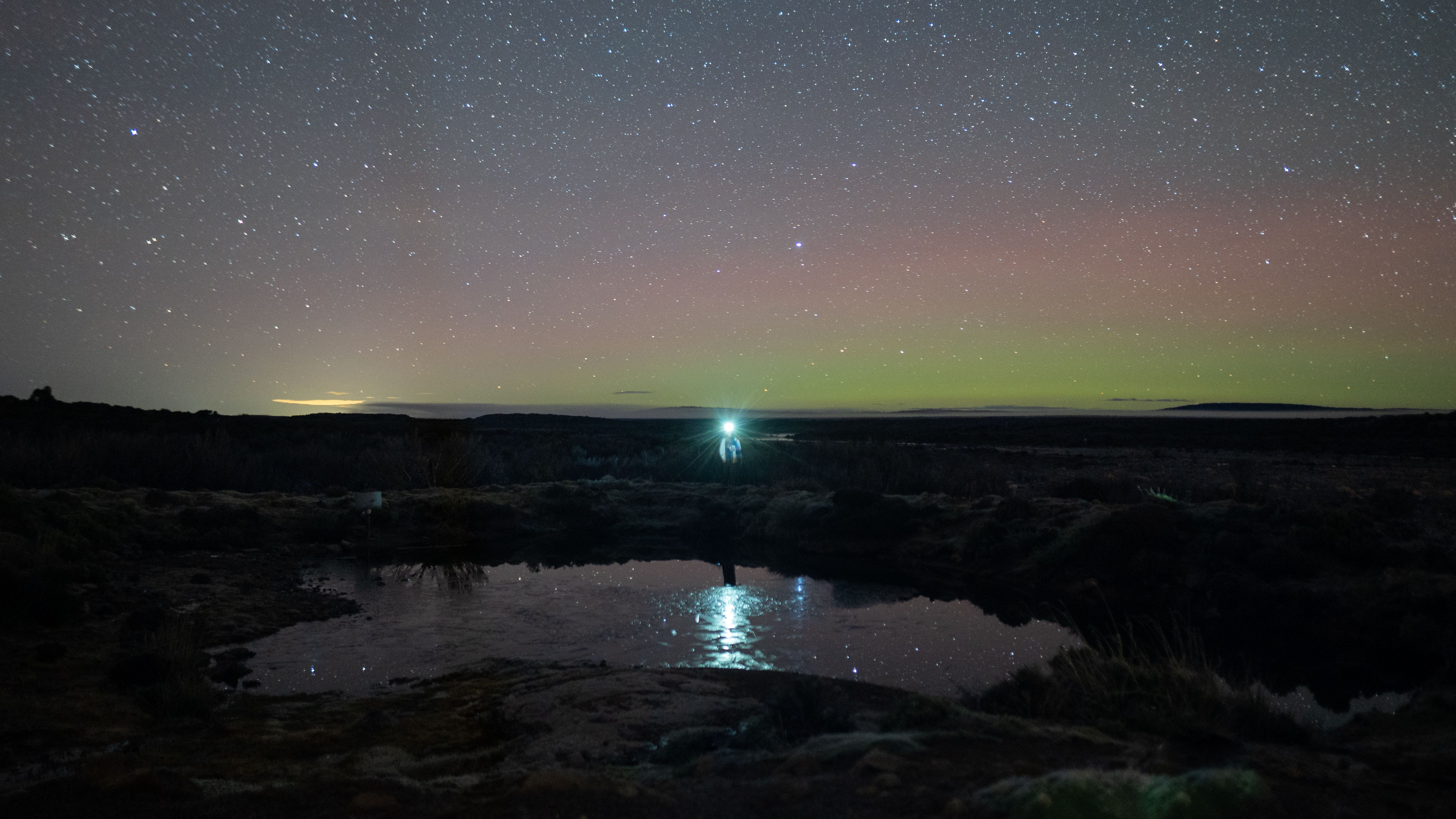 Person with a head torch shinning at the camera with the Aurora Australis lighting up the night sky green and pink.