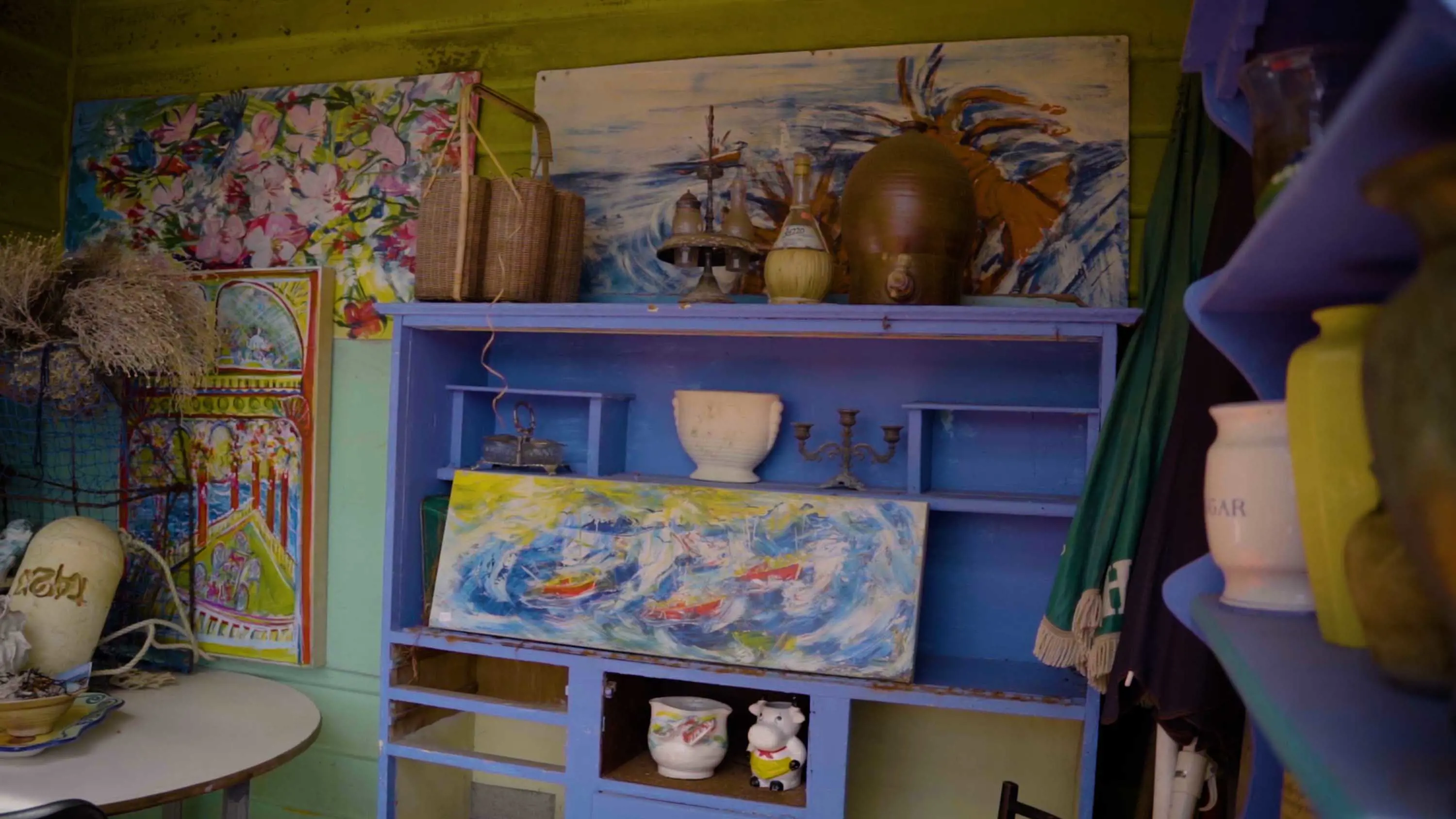 A painting of a wild sea with boats sits on a the shelf of a lavendar coloured cabinet.