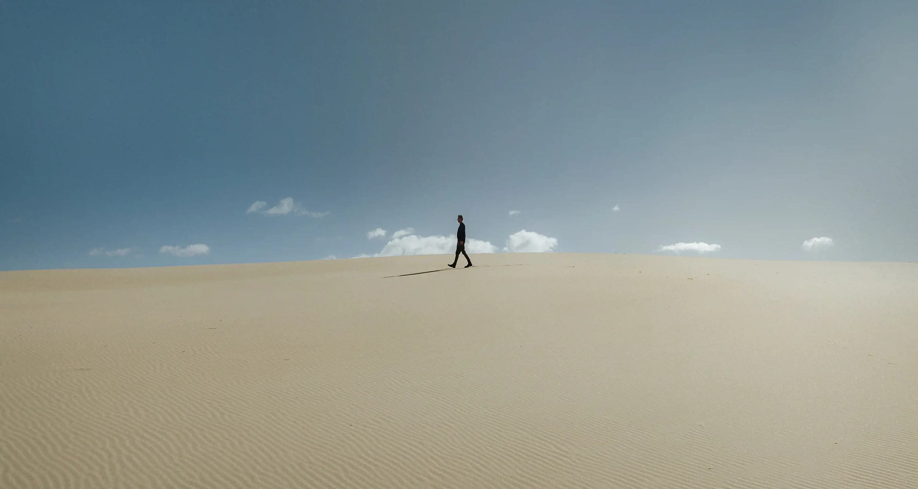 A man walks along the top of a large sand dune on a almost cloudless, sunny day.