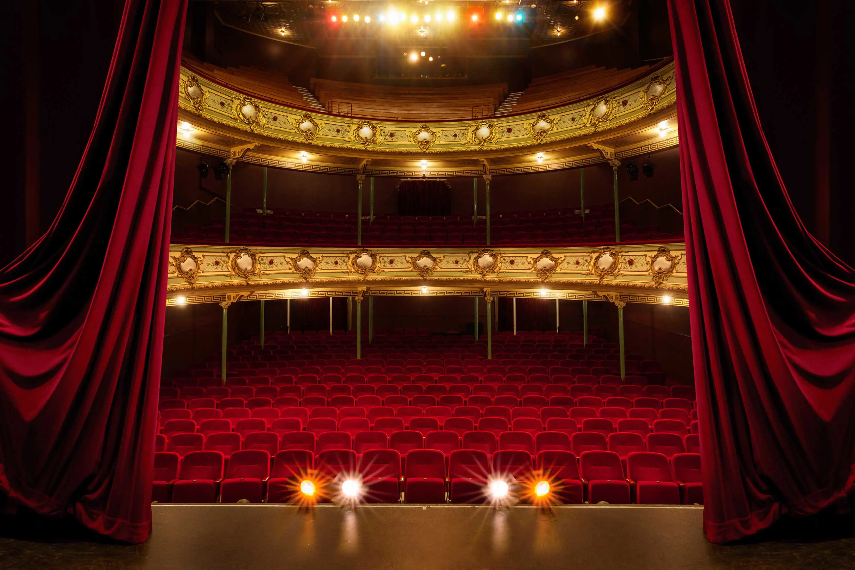 A the stage of traditional theatre in front of red velvet seats on two tiers is framed by enormous red curtains.