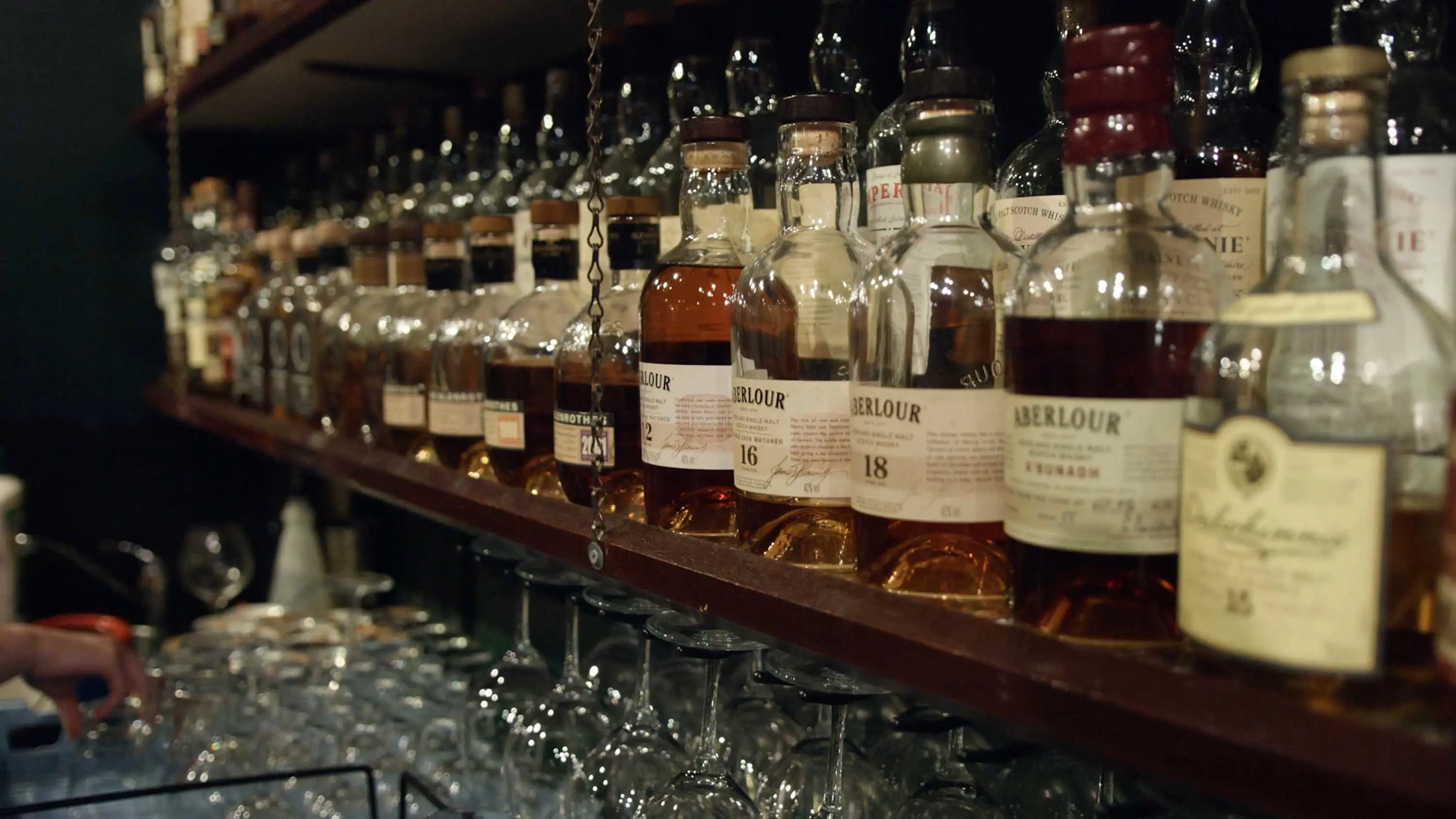 A line of whisky bottles of various sizes sitting on a shelf above glassware.